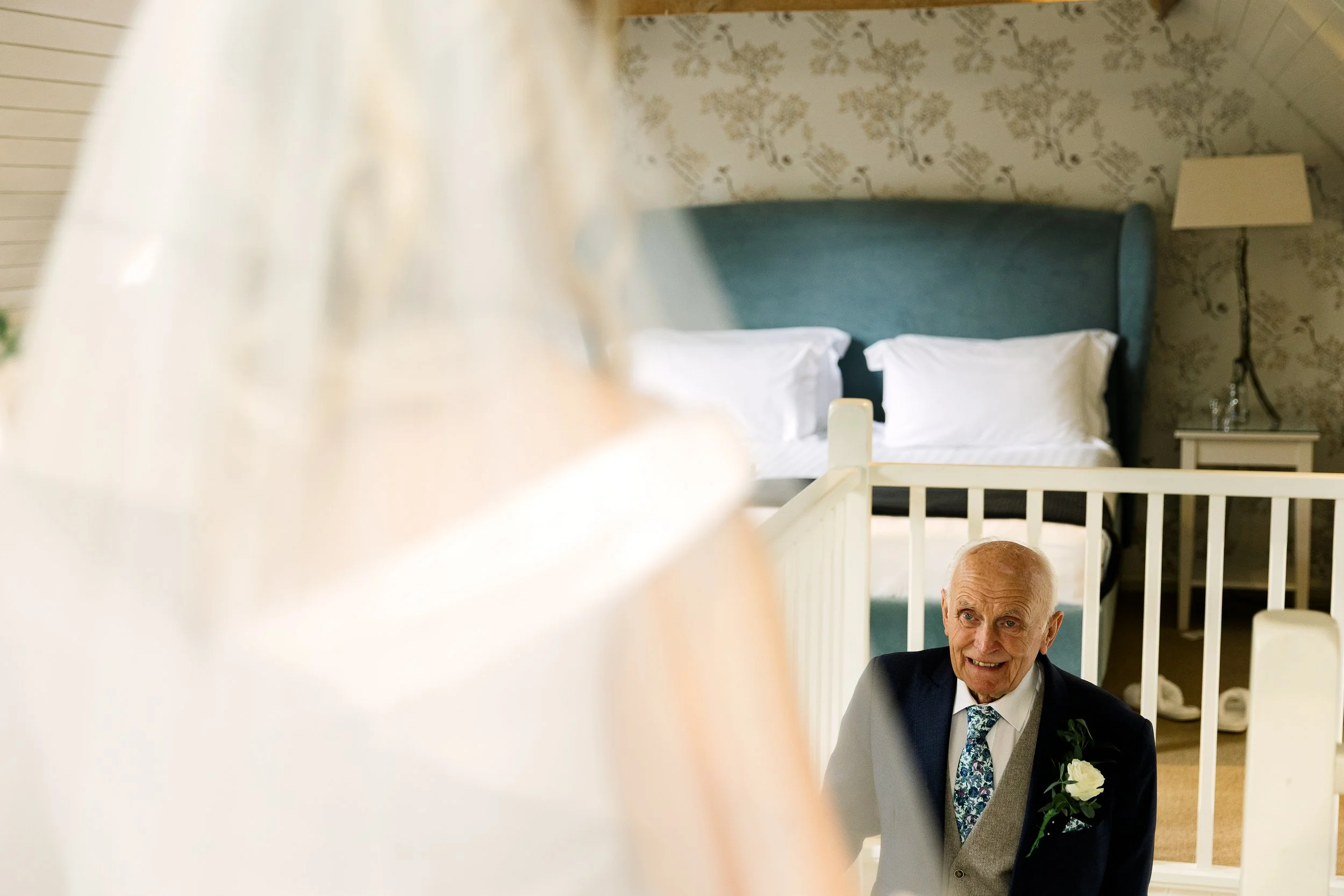 An elderly man in a suit with a boutonniere, smiling, possibly at a wedding or special event, seen through a blurred foreground of a woman in white.