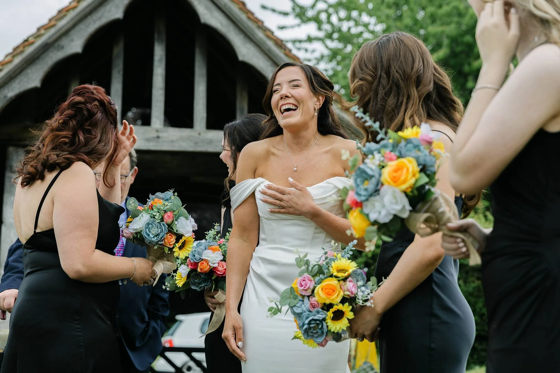 A woman in a white off-shoulder dress is laughing and touching her chest surrounded by women holding bouquets of colorful flowers during a wedding celebration outdoors.