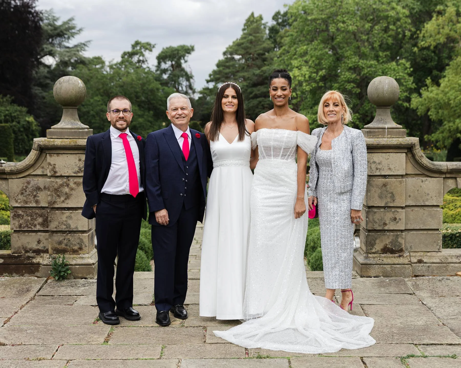 Group of five people, including a bride in a white wedding dress, posing outdoors on a stone terrace with greenery and trees in the background.