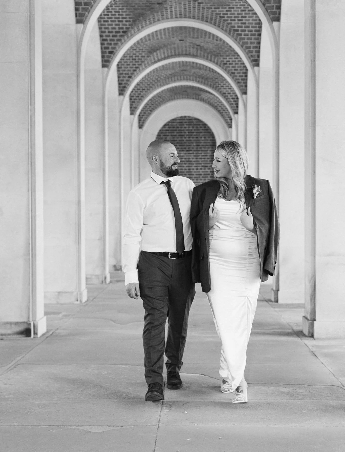 A black-and-white photo of a couple walking through an arched walkway; the man, with a beard, in a dress shirt and tie, looks at the woman, who is in a long white dress and a jacket draped over her shoulders, smiling at each other.