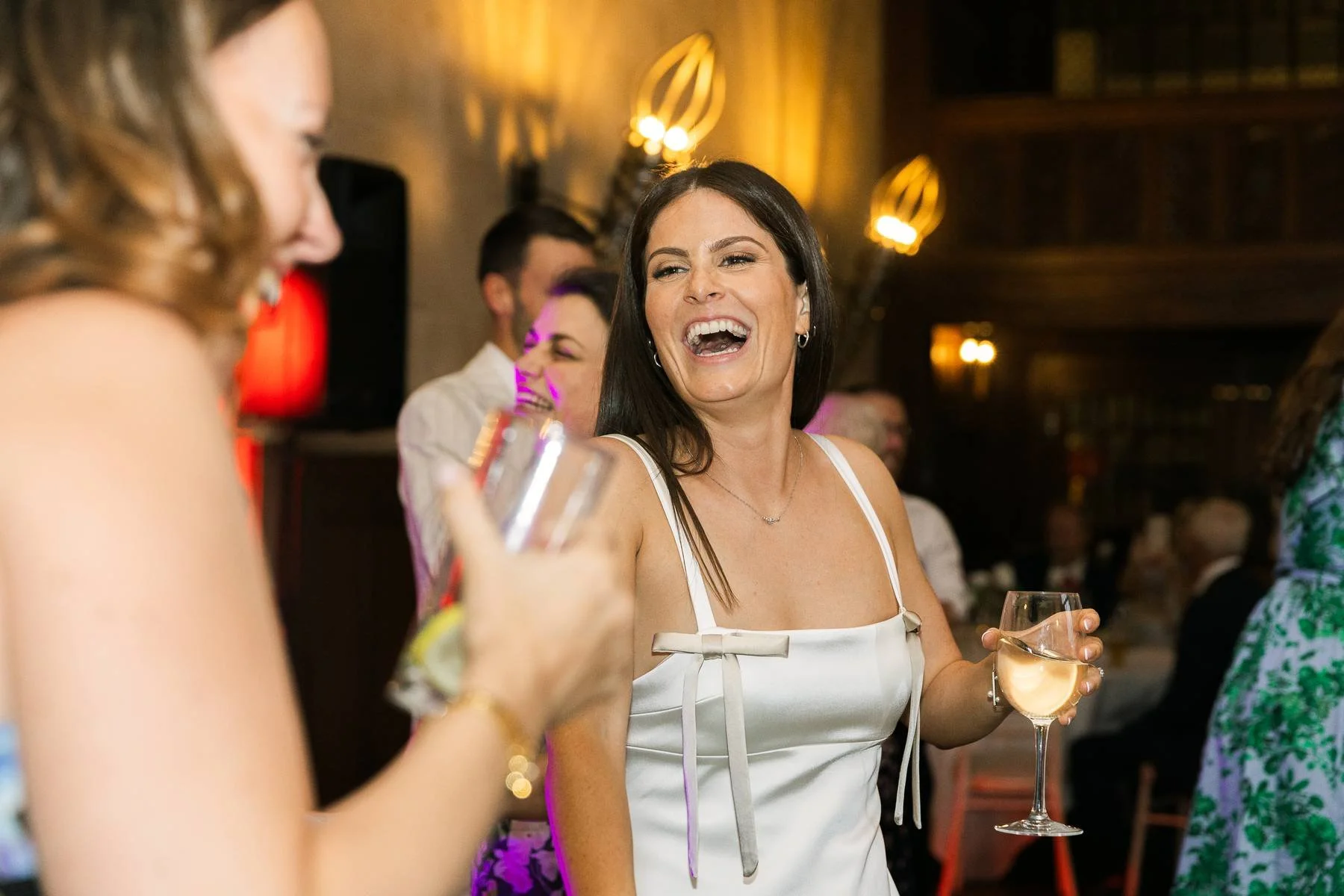 A woman with dark hair wearing a white dress and holding a glass of white wine, laughing at a social event or party.