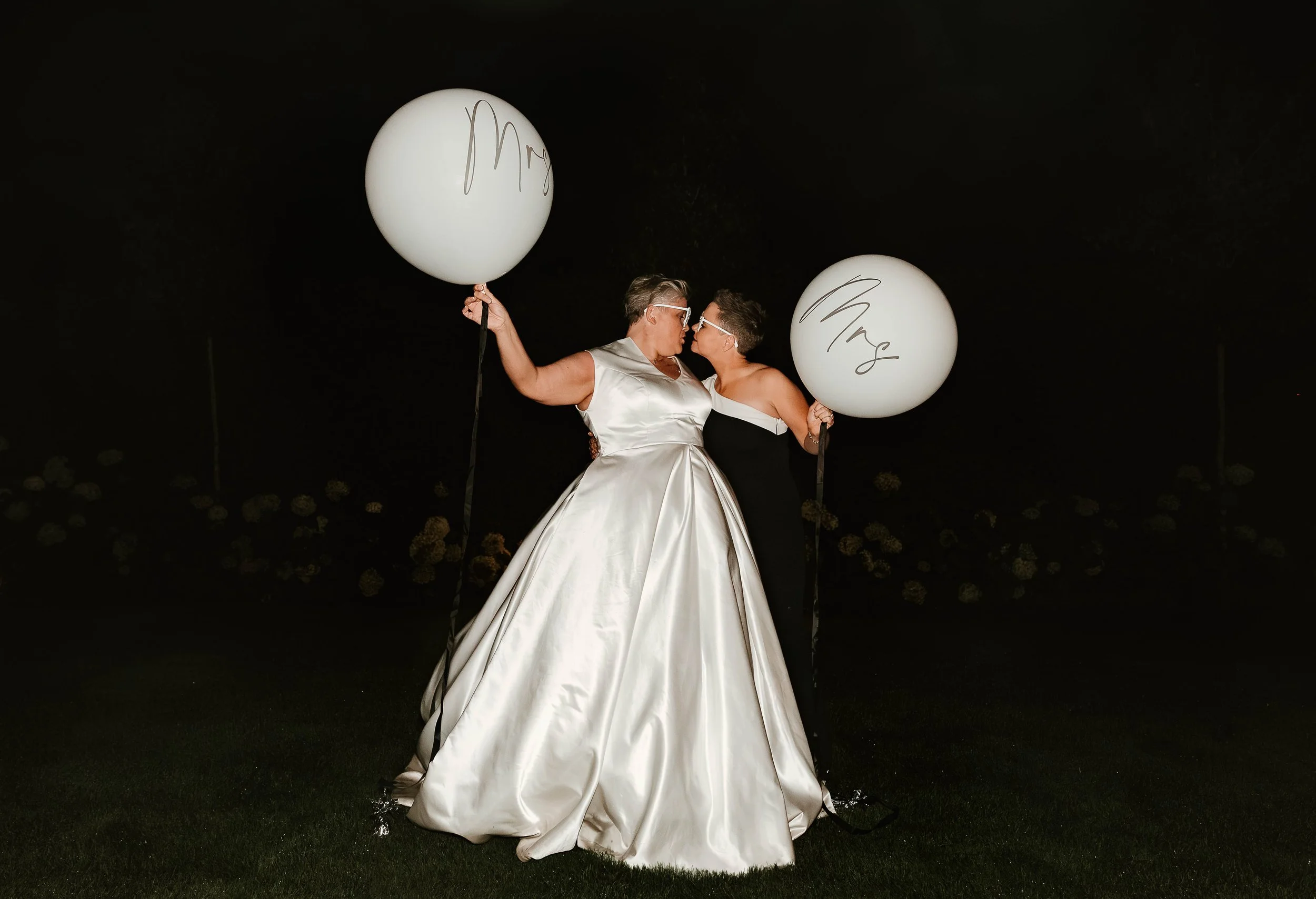 Two women, one in a white satin dress and the other in a black dress, stand close together holding white balloons with 'Mrs' written on them, at night outdoors.