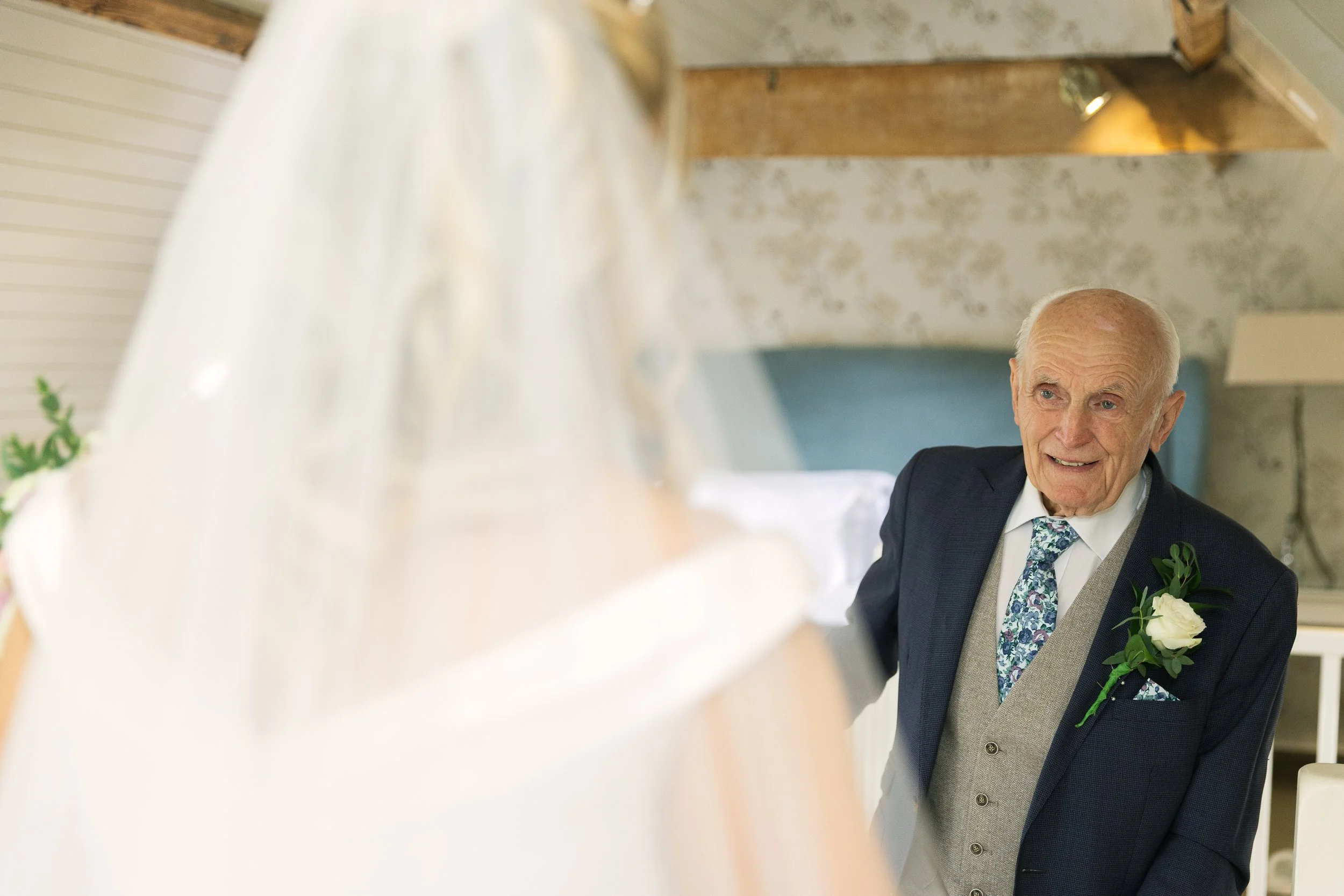 An elderly man in a suit with a flower pinned to his lapel, smiling and looking towards a bride wearing a wedding veil and dress.