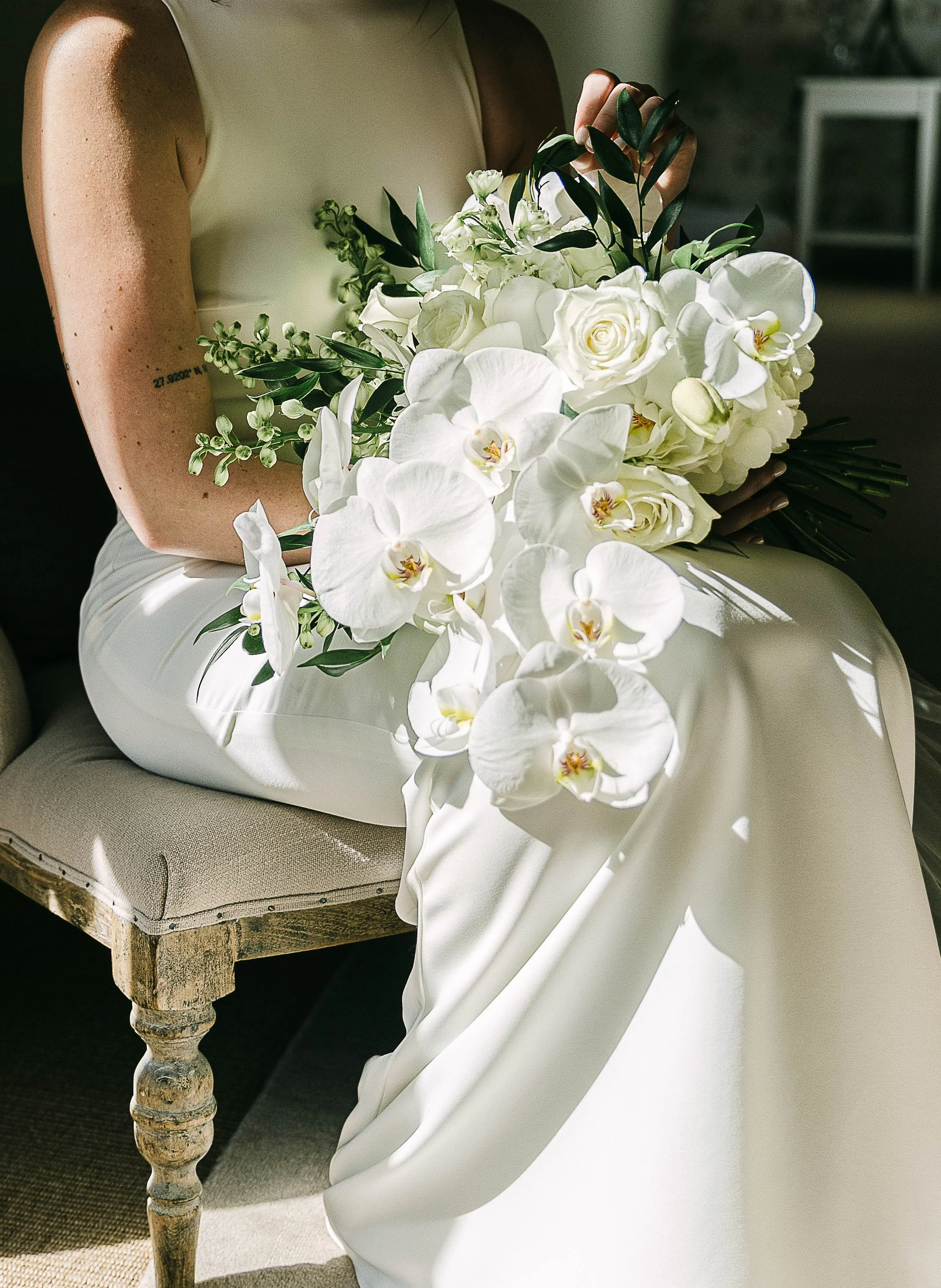 A person dressed in white holding a large bouquet of white flowers, including roses and orchids, while sitting on a vintage wooden chair with a beige cushion.