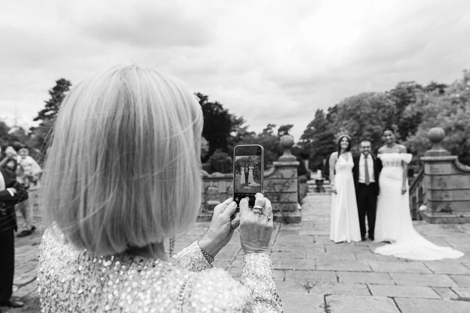 A woman with blond hair taking a photo of two women and a man standing outdoors on a cloudy day.