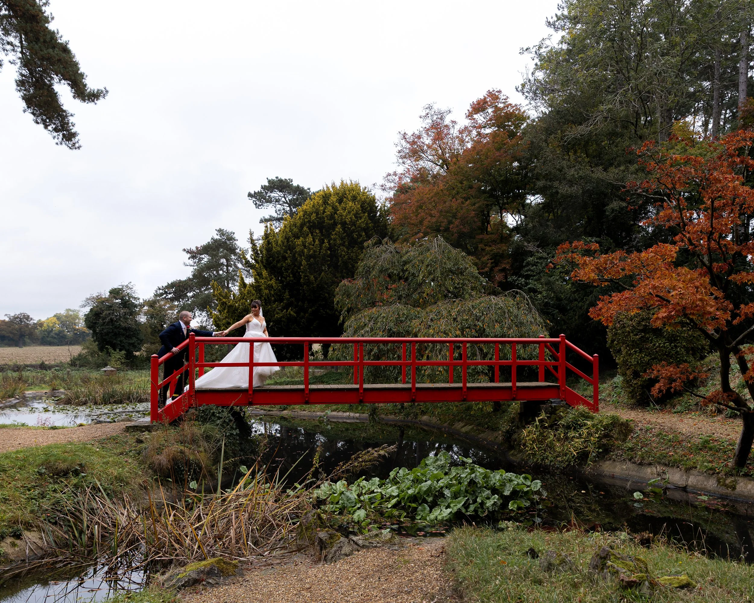 A bride and groom holding hands on a red footbridge in a scenic park with autumn foliage