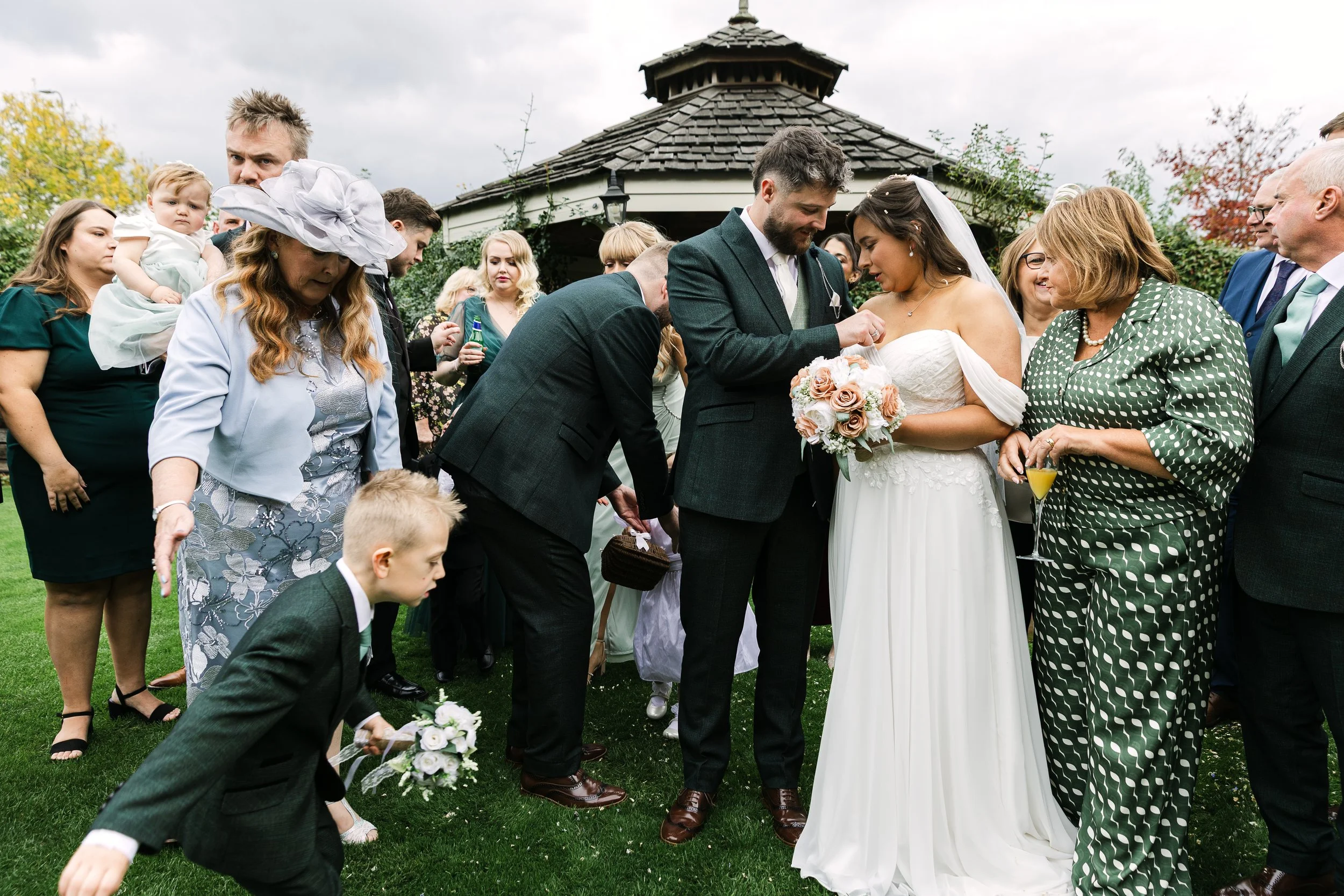 A wedding celebration outdoors with a bride and groom in the center, surrounded by family and friends. The bride wears a white dress and holds a bouquet, while the groom wears a suit. People are standing on green grass in front of a wooden structure with trees and a cloudy sky in the background.