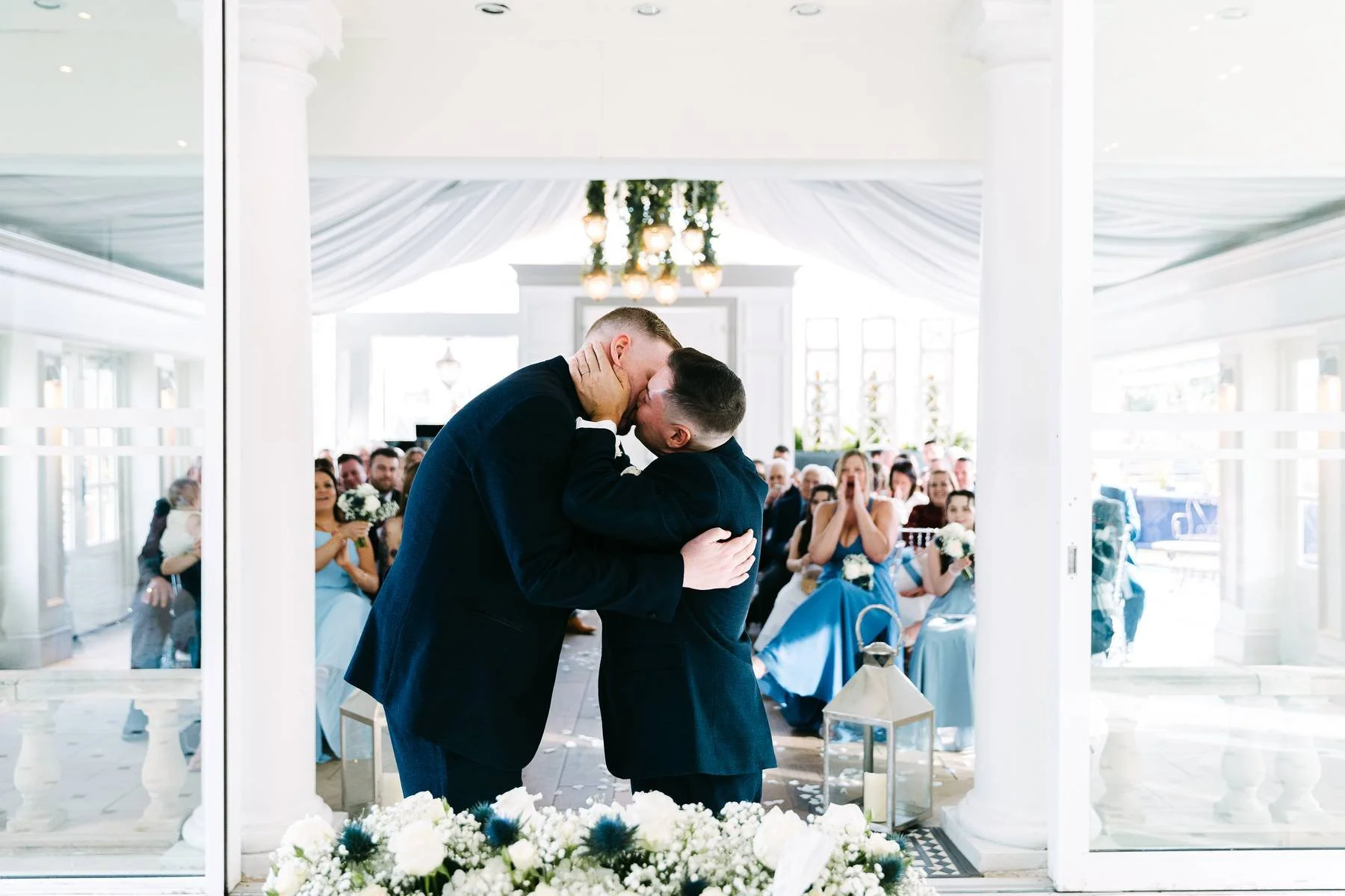 Two men share a kiss during a wedding ceremony, with guests sitting in the background and floral decorations around them.