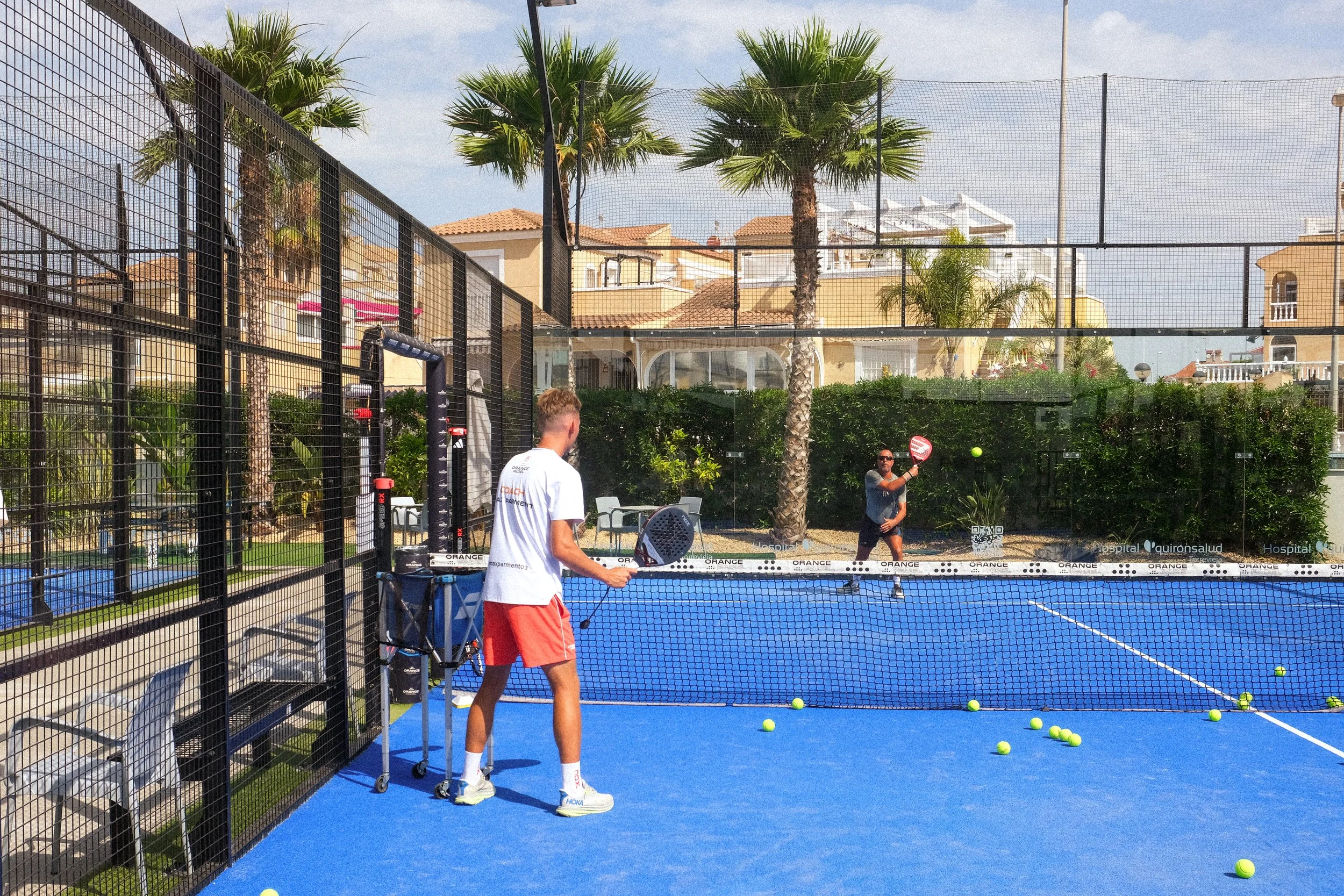 Two men playing padel on a blue court outdoors surrounded by a black safety fence, with palm trees and residential buildings in the background, and tennis balls scattered on the court.
