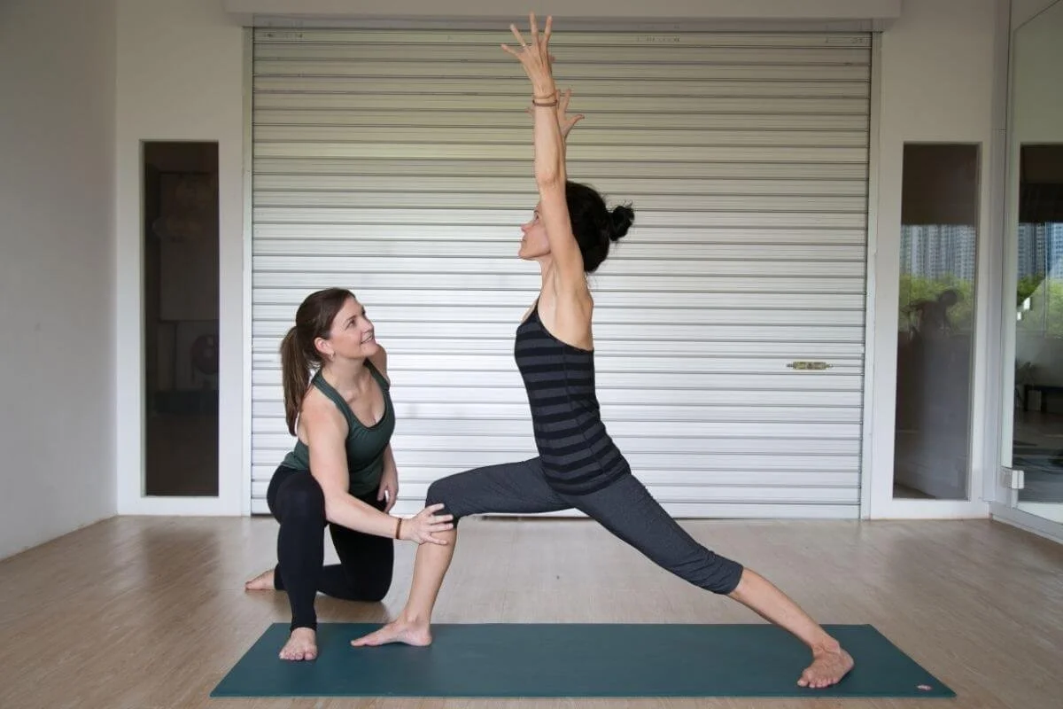 Yoga teacher observing and assisting a student's knee alignment in Warrior 1 to support a student with pain