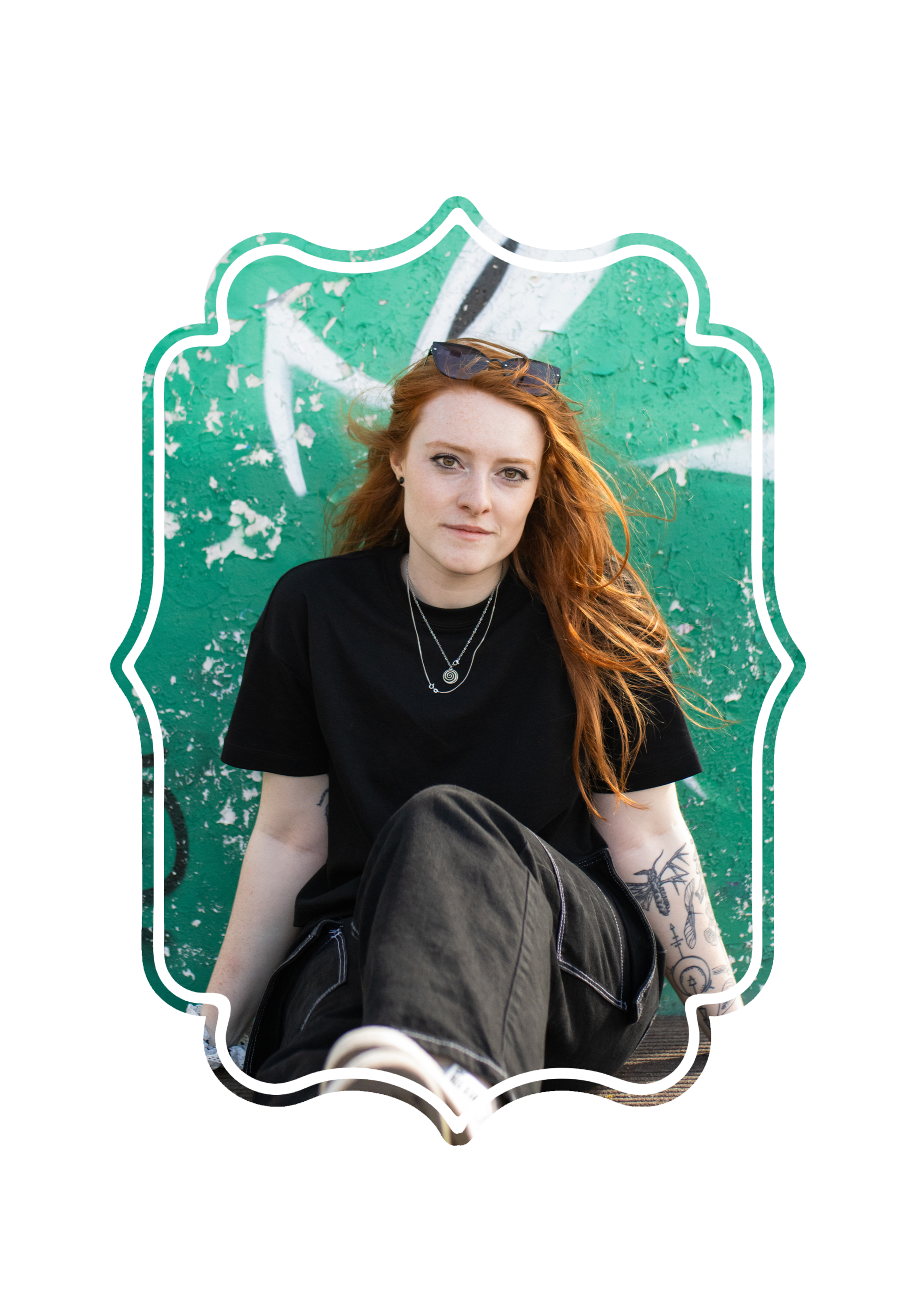 A young woman with red hair and sunglasses on her head, wearing a black shirt and silver jewelry, sitting in front of a green wall with white graffiti, with a tattooed arm visible.
