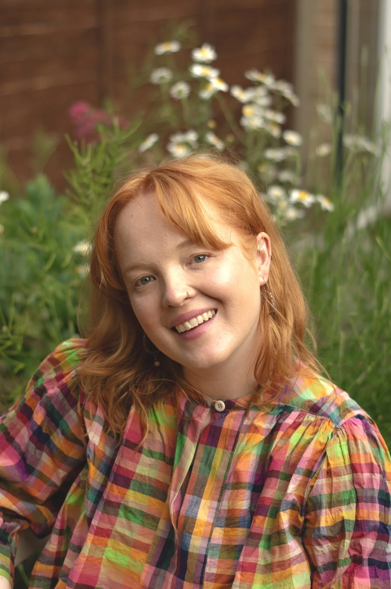 A woman with red hair and a blue blouse sitting on a gray couch in a modern living room, smiling at the camera, with colorful abstract art on the wall behind her and a yellow lamp in the background.