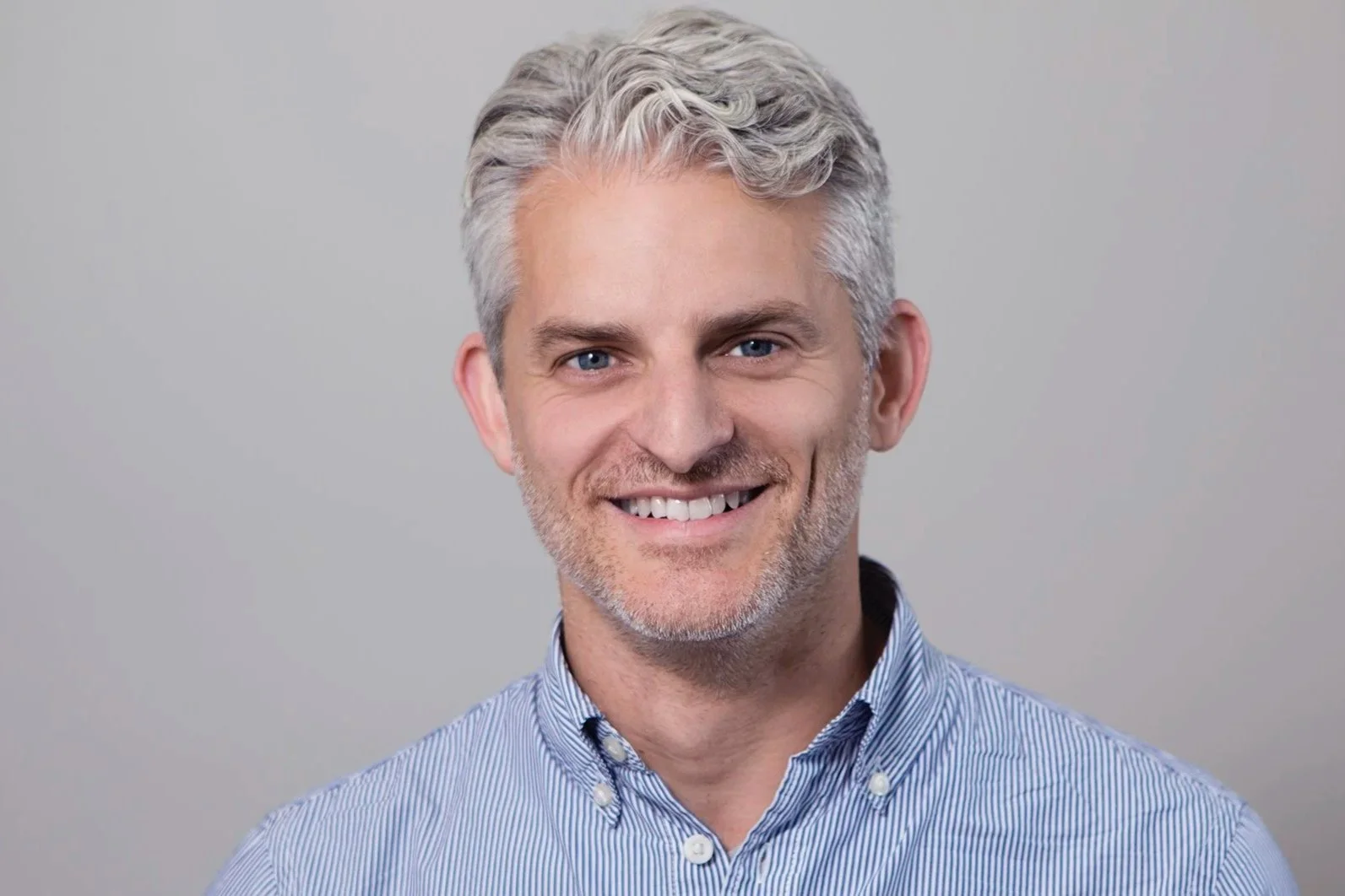 Man with short, wavy white hair smiling directly at the camera with a light, blank background. 