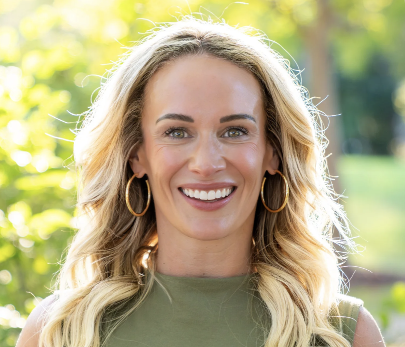 Young woman with wavy blond hair smiling and looking into the camera with sunlight and foliage in the background. 