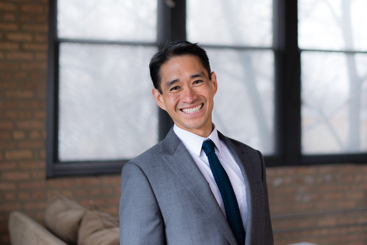 Voltaire Santos Miran in a suit and knit tie standing indoors near a window with brick wall background.