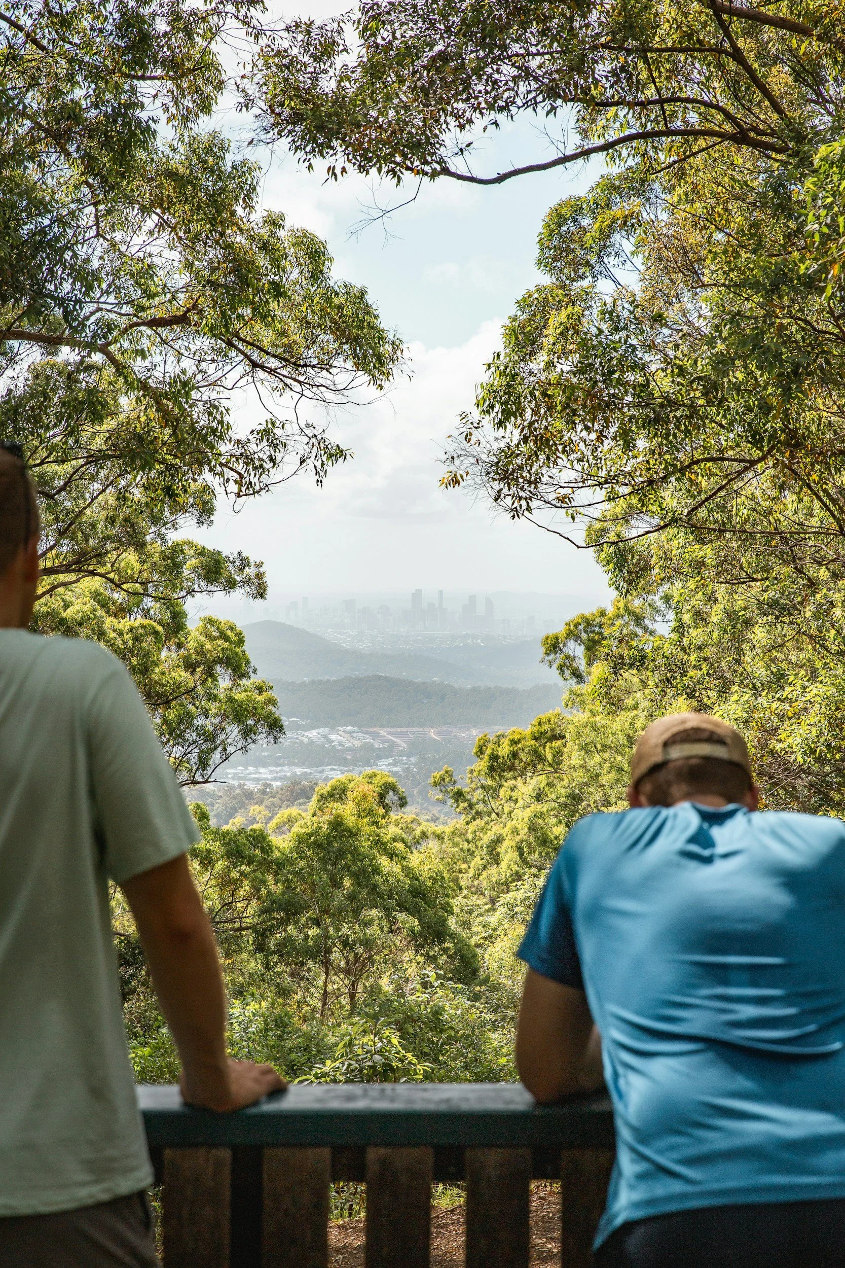 Two men standing and sitting on a wooden railing on a mountain overlook, viewing a city skyline in the distance through a forested area.