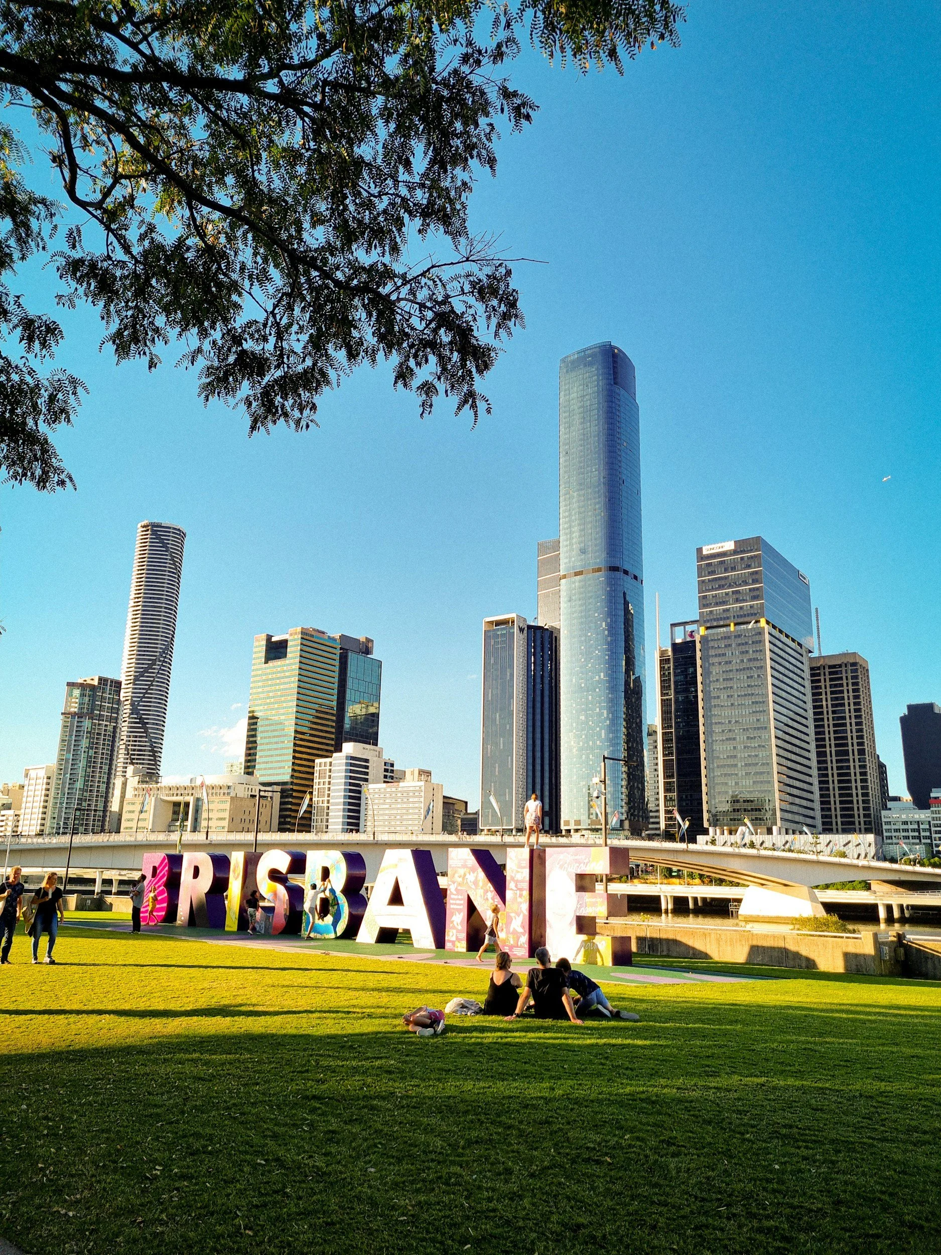 People relaxing on a grassy area near a large, colorful 'BRISBANE' sign with a city skyline and tall skyscrapers in the background.