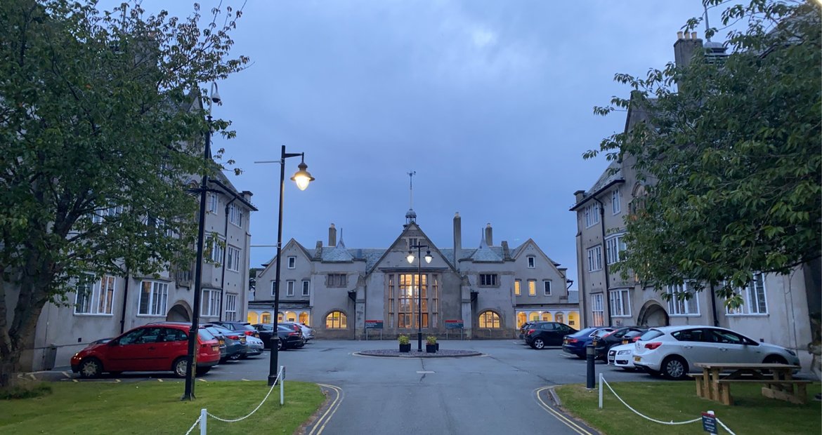 A large, historic building resembling a hotel or an apartment complex, with cars parked in front, trees on either side, and streetlights illuminated at dusk.