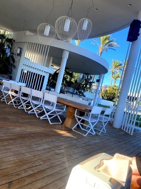 Outdoor patio area with white chairs and tables, hanging pendant lights, and a modern building with palm trees in the background.