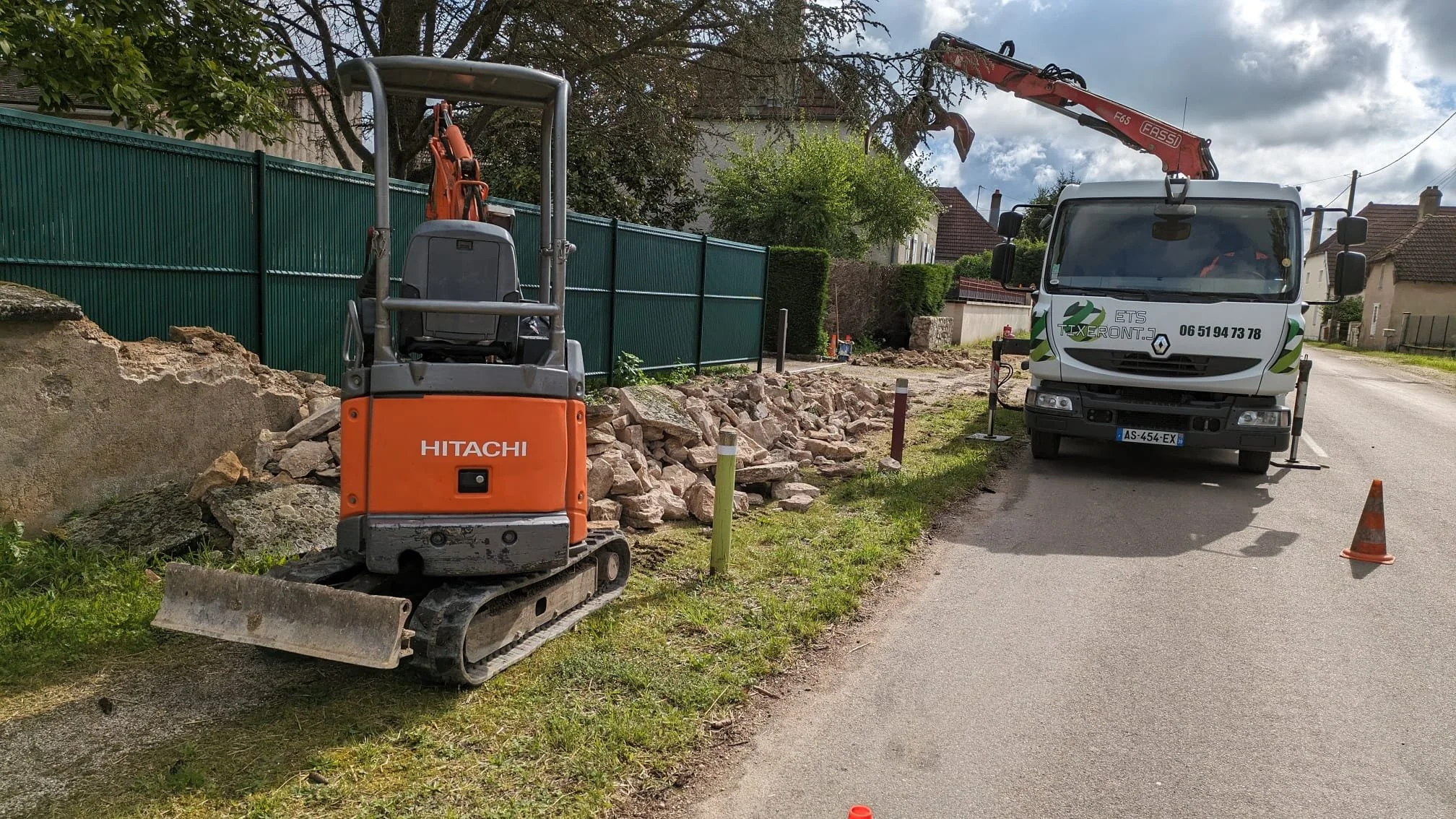 Travaux de voirie avec une mini-pelle Hitachi orange et un camion avec nacelle élévatrice, entourés de cônes de signalisation, dans une rue résidentielle.