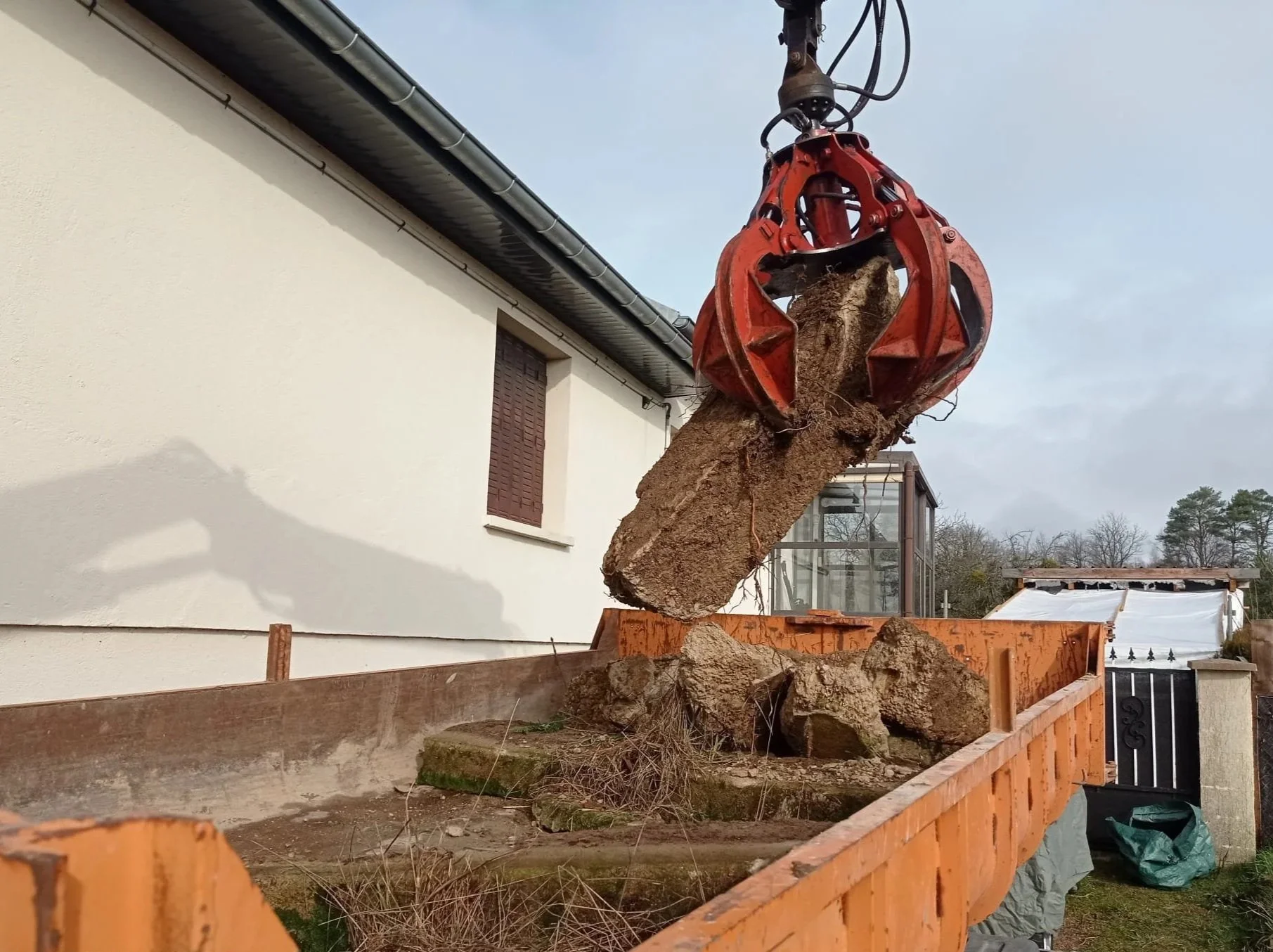 Une grue de construction décharge un gros bloc de pierre dans une benne en bois. La scène se déroule dans une cour résidentielle avec une maison blanche en arrière-plan.