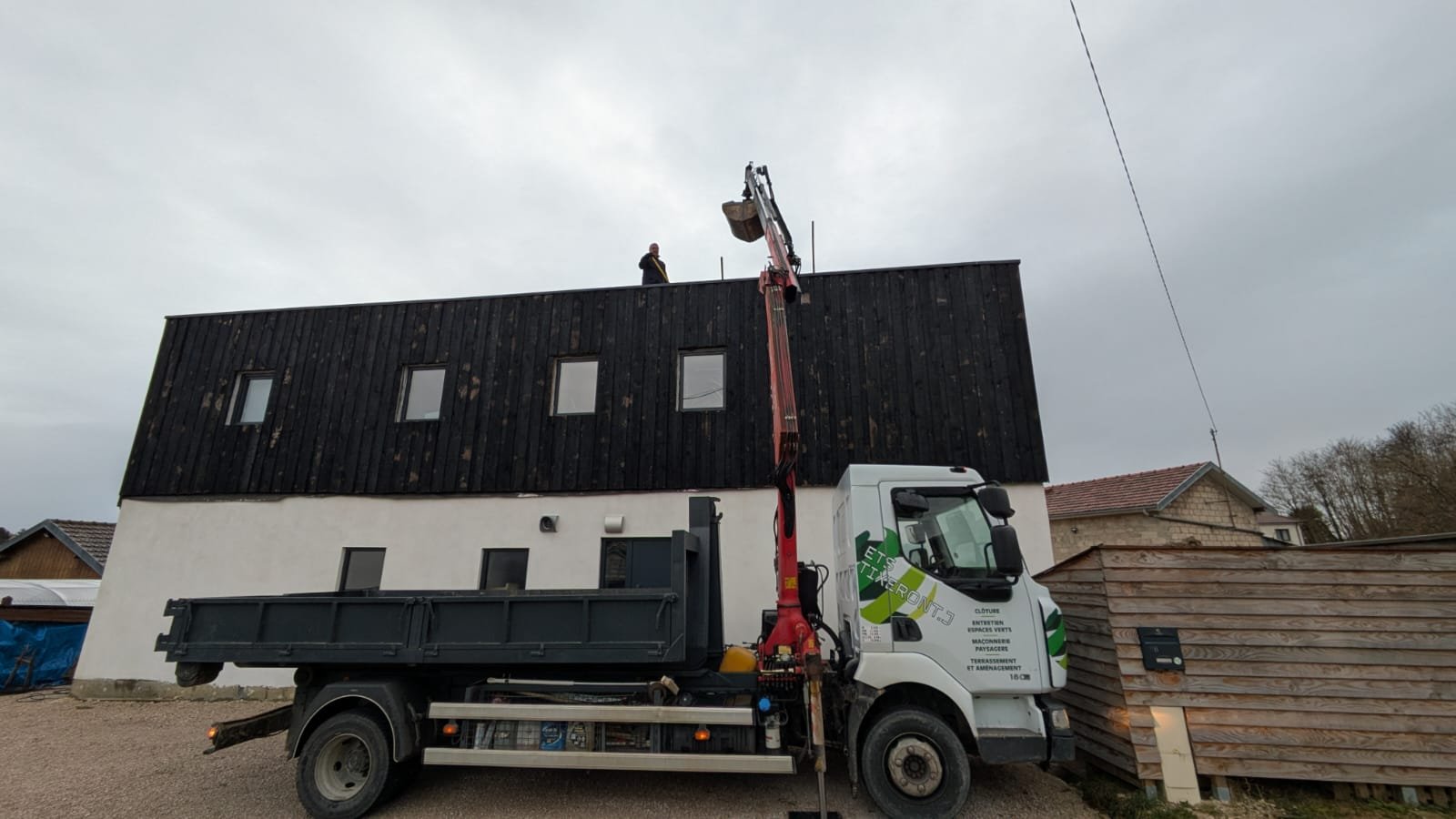 Ouvrier utilisant une grue pour travailler sur le toit d'une maison moderne à deux étages, avec de petites fenêtres, un ciel nuageux et des maisons voisines en arrière-plan.