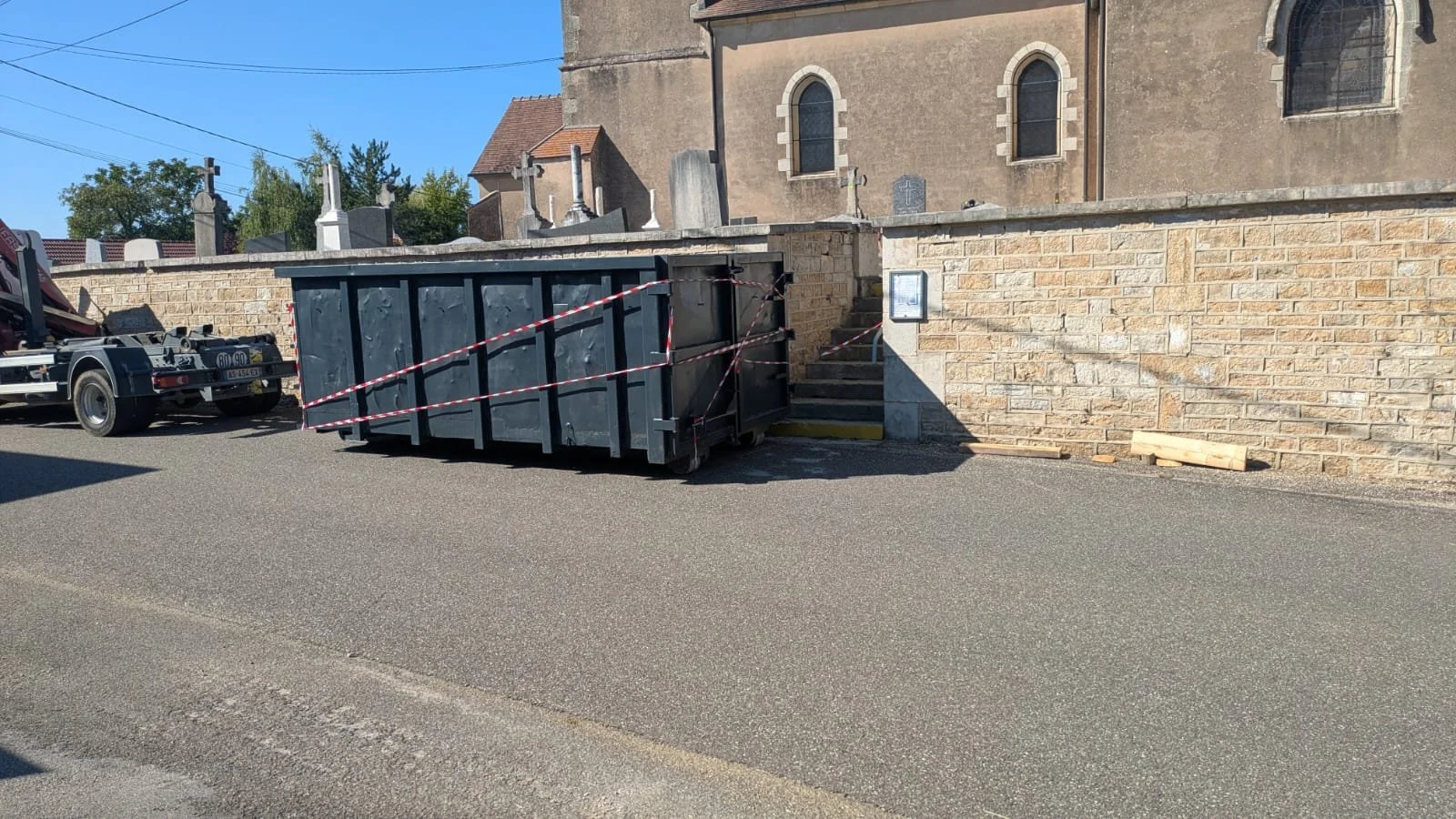 Une benne à ordures noire bloquée par une bande de ruban rouge et blanc, placée contre un mur en pierre d'une vieille église ou bâtiment religieux, avec un petit escalier à côté et une plaque d'information. Le trottoir est asphalté et une voiture est visible à gauche. Le ciel est bleu et il fait beau.