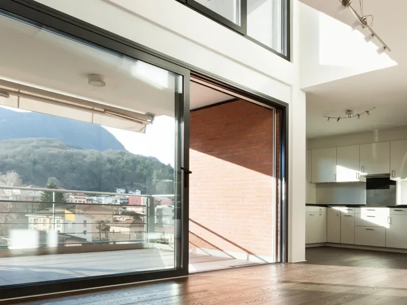Sala moderna con pisos de madera, grandes ventanas corredizas que llevan a un balcón con vista a una ciudad y montañas, cocina blanca en el fondo con electrodomésticos integrados.
