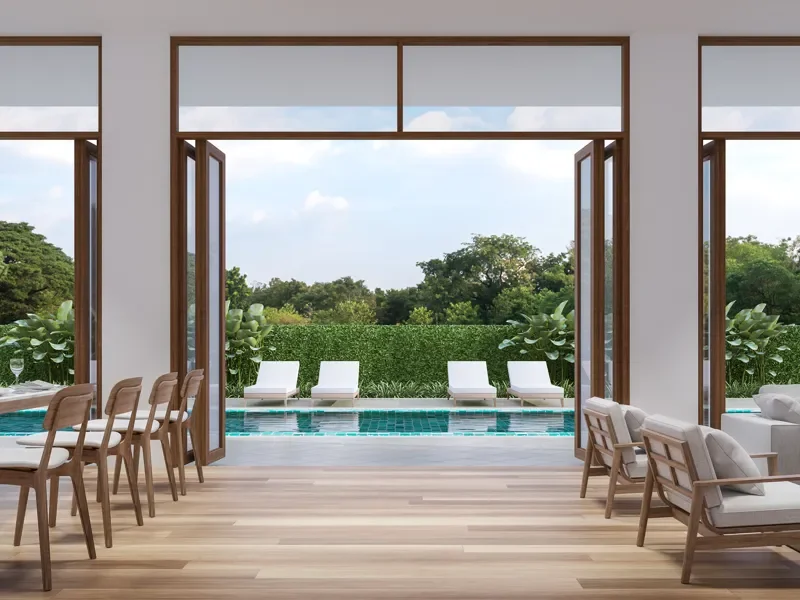 Interior de una casa con vista a una piscina, con sillones y sillas de madera, árboles y cielo despejado en el exterior.