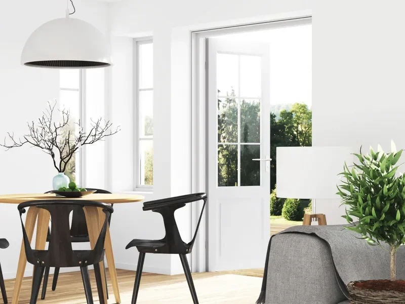 Interior de una sala moderna con puertas de vidrio abiertas, una lámpara blanca colgante, una mesa redonda de madera con sillas negras, una planta en una esquina y mucha luz natural.