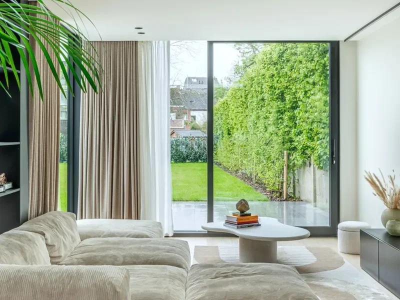 Sala moderna con sofá beige, mesa de centro blanca, cortinas beige y blancas, vista a un jardín con césped y árboles a través de ventanas grandes.