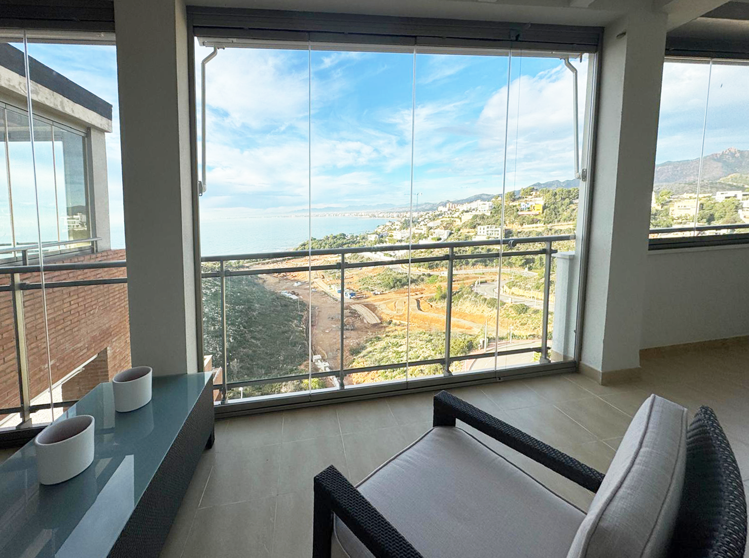 Vista panorámica de un balcón con vista al mar y a la ciudad desde un apartamento moderno, con una silla y una mesa decorativa con macetas.