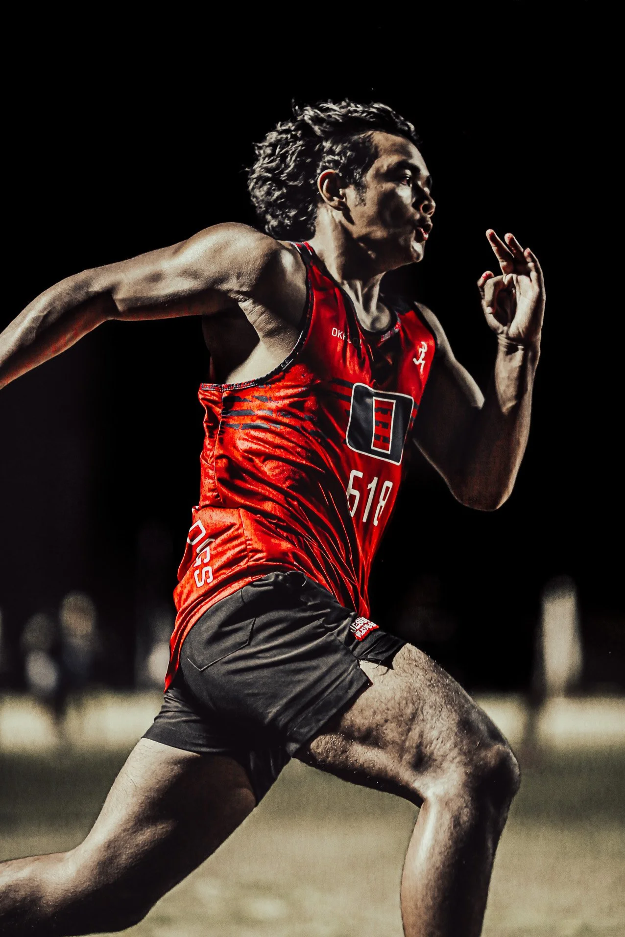 A male athlete running on a sports field at night, wearing a red and black sports uniform with the number 518, with dark hair and a focused expression.