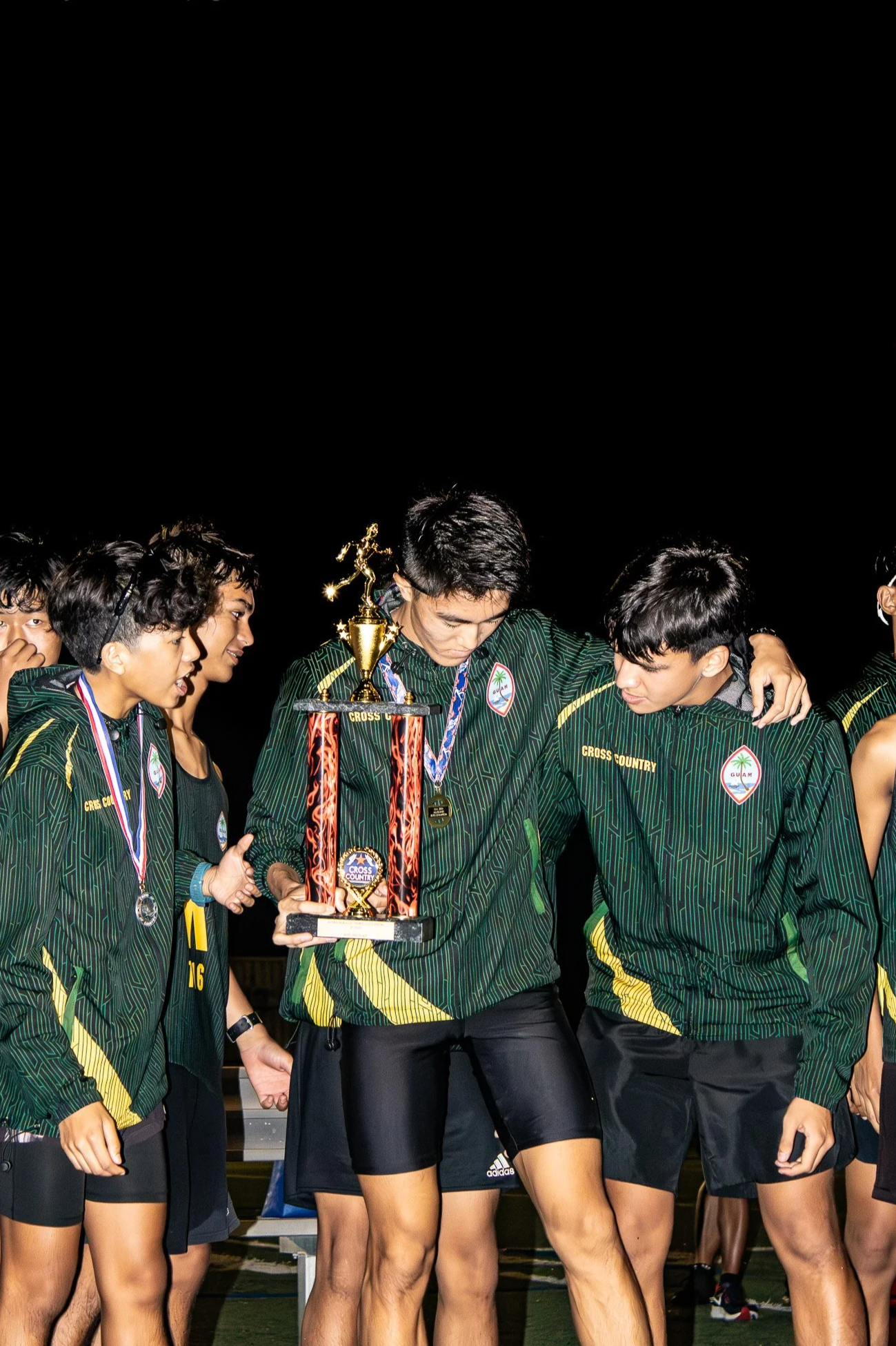 A group of young athletes in green and black uniforms celebrating at night, with one holding a large trophy and wearing a medal.