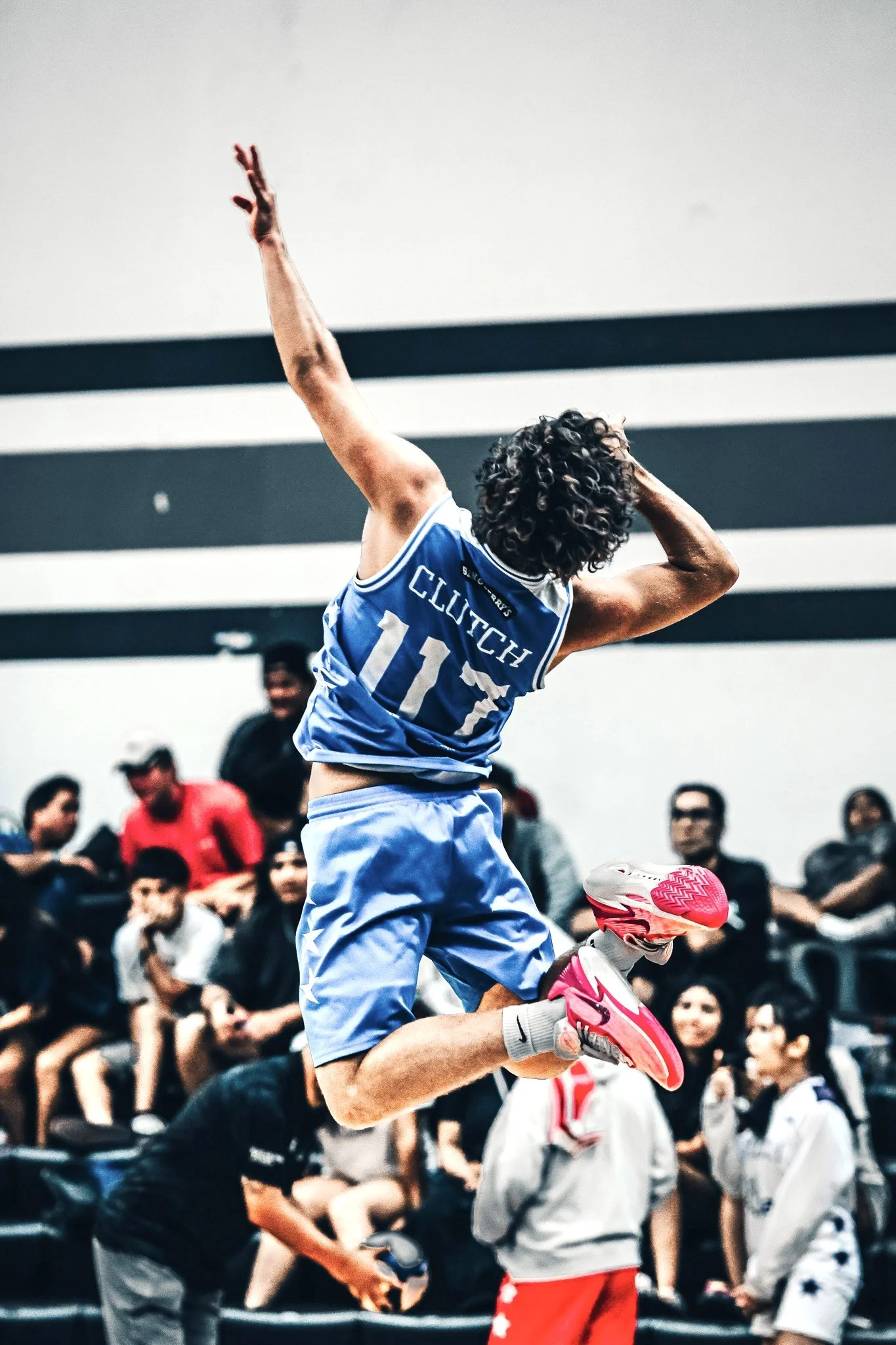 A basketball player wearing a blue jersey with the number 11 and the name 'Clutch' is jumping mid-air during a game, with spectators watching from the background.