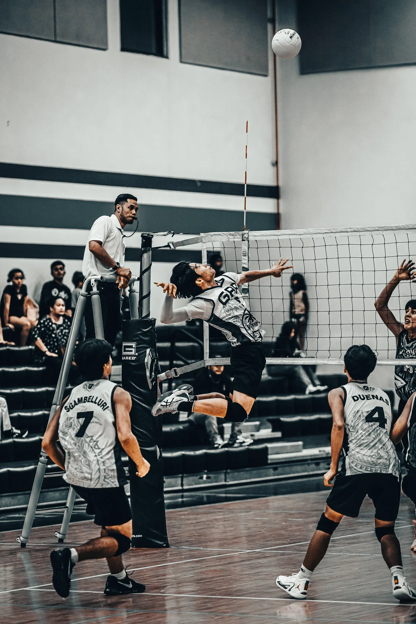 A volleyball game in action with players jumping near the net and an official overseeing the game.