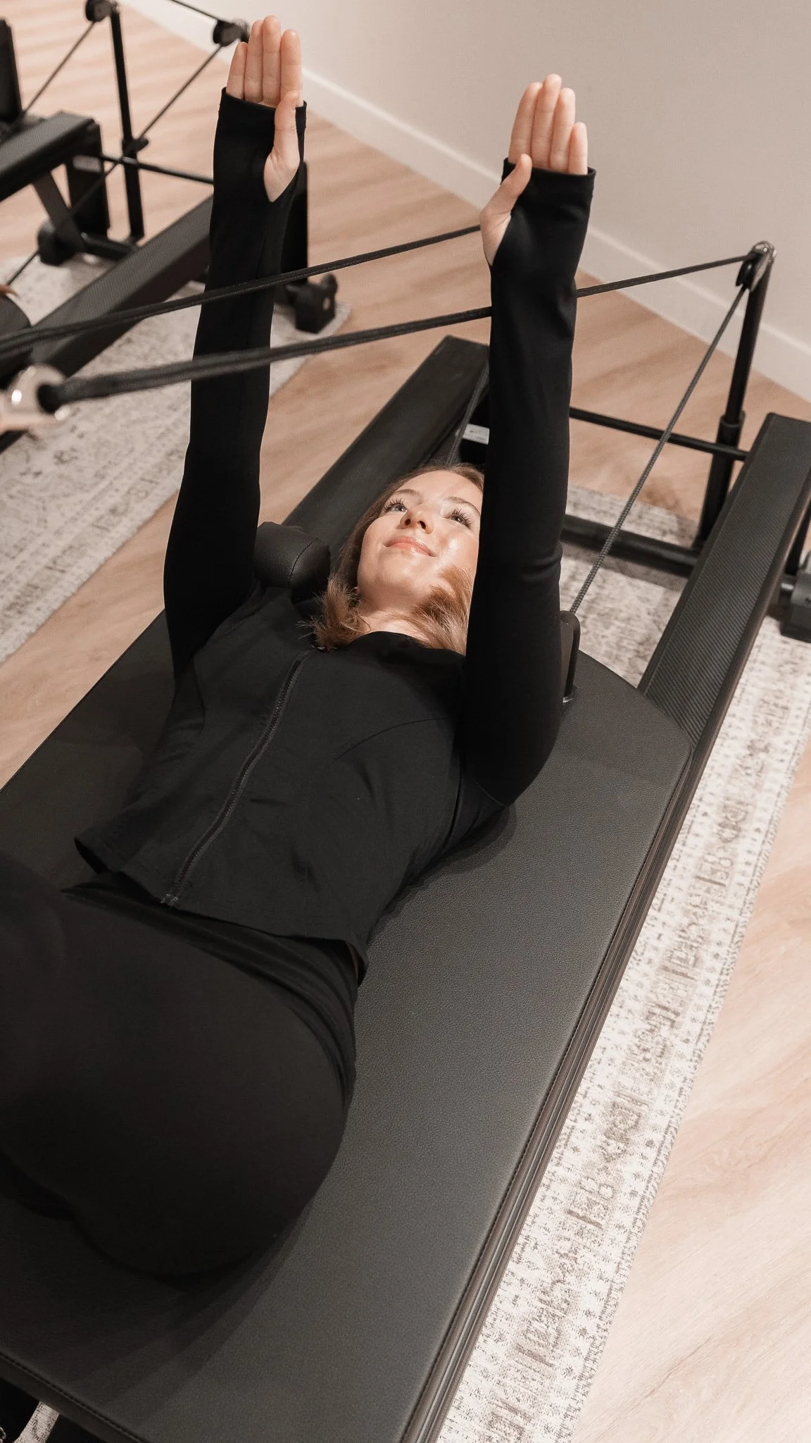 Femme allongée sur un appareil de Pilates ou de yoga, portant des vêtements de sport noirs, les bras levés, dans une salle d'entraînement intérieure.