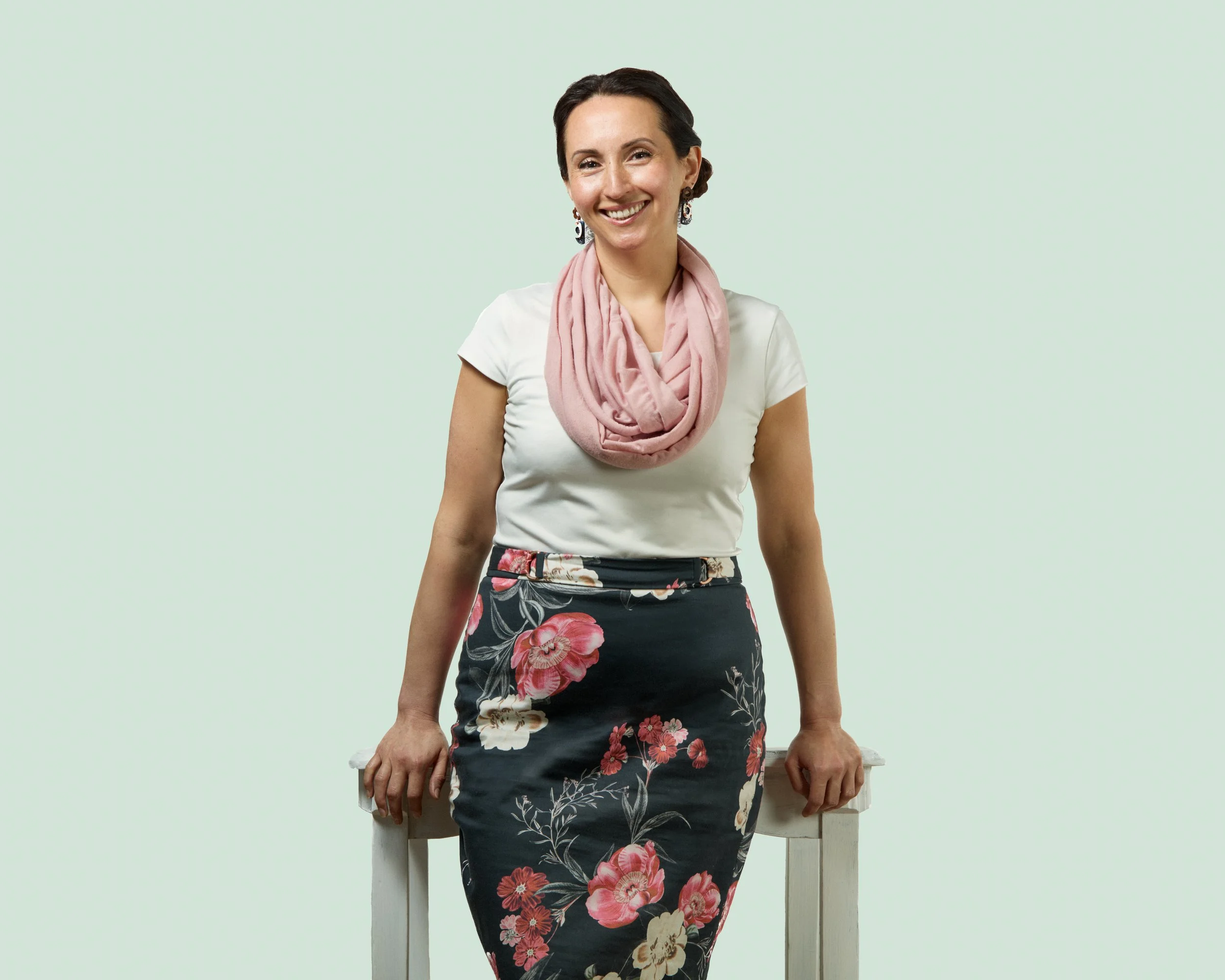 Young woman with dark hair wearing a white t-shirt, pink scarf, and a floral skirt, leaning on a white table against a light green background, smiling.