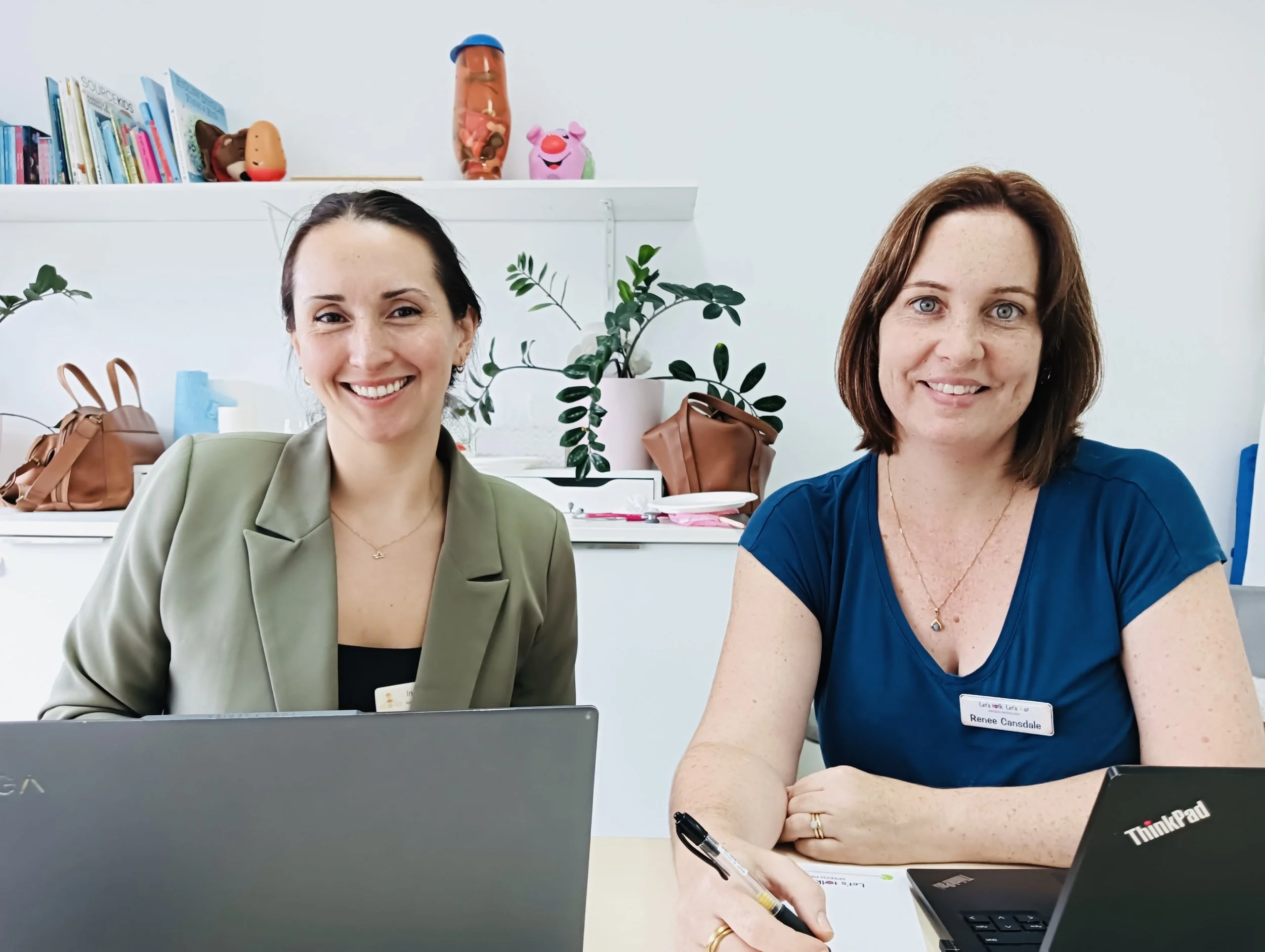 Two women sitting at a desk, smiling at the camera, with laptops and office supplies in a bright, organized office.