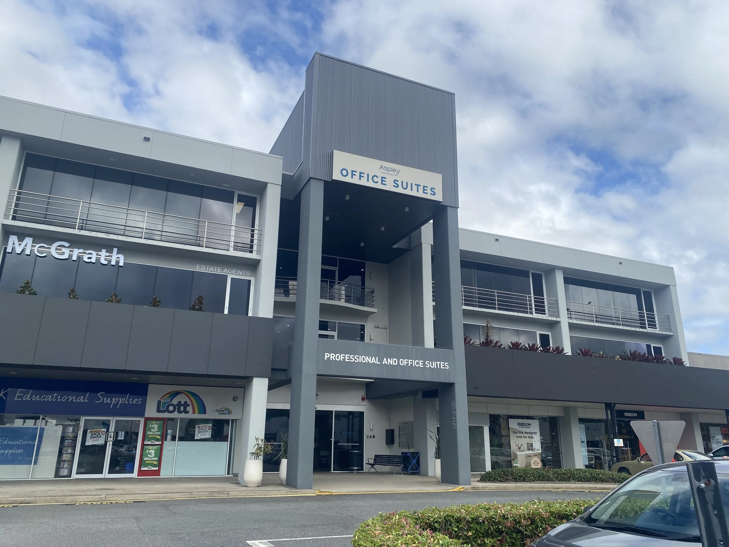 A modern commercial building with signs for office suites, estate agents, and educational supplies, blue sky with clouds.