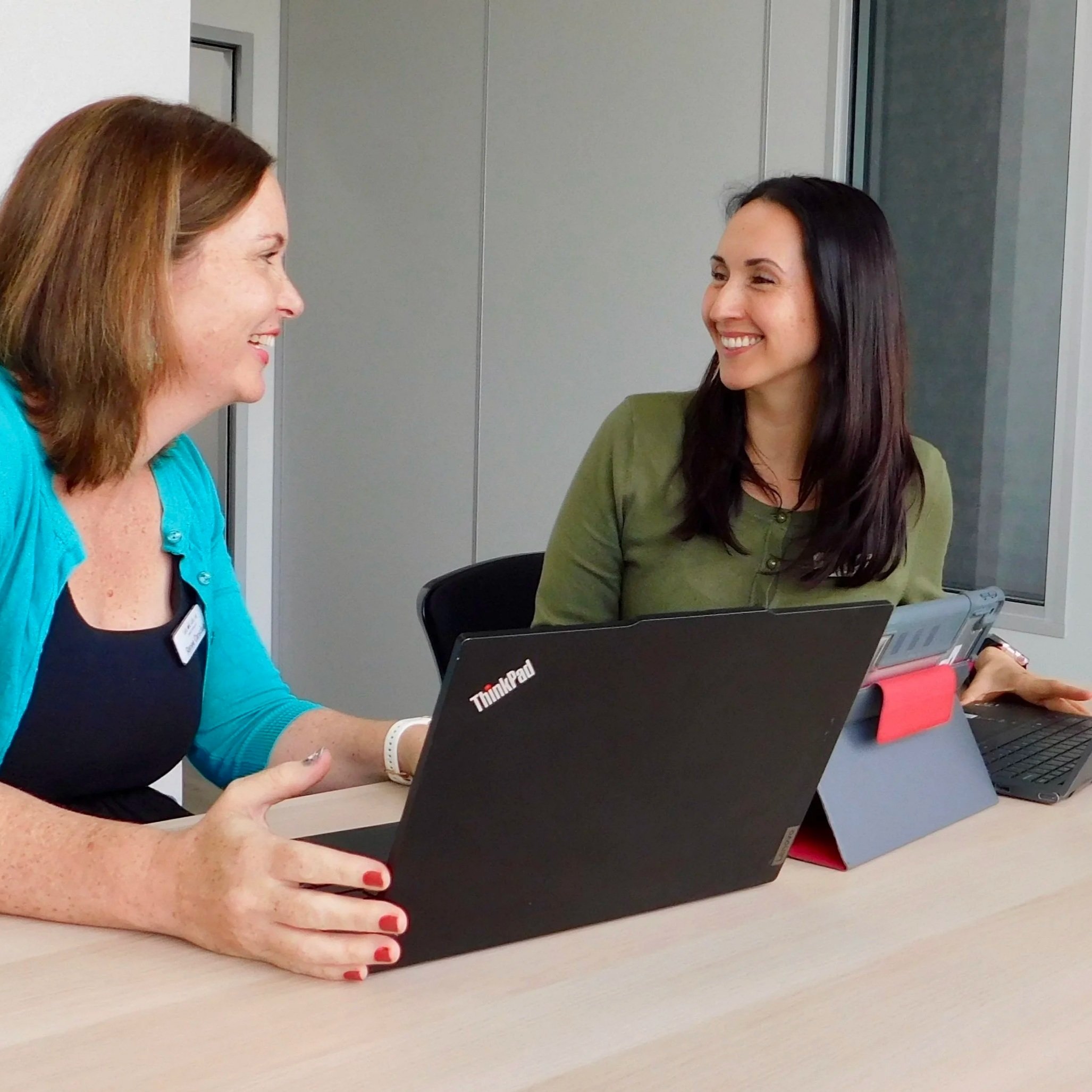 Two women sitting at a table, smiling and engaged in a conversation, with laptops in front of them in a modern office setting.