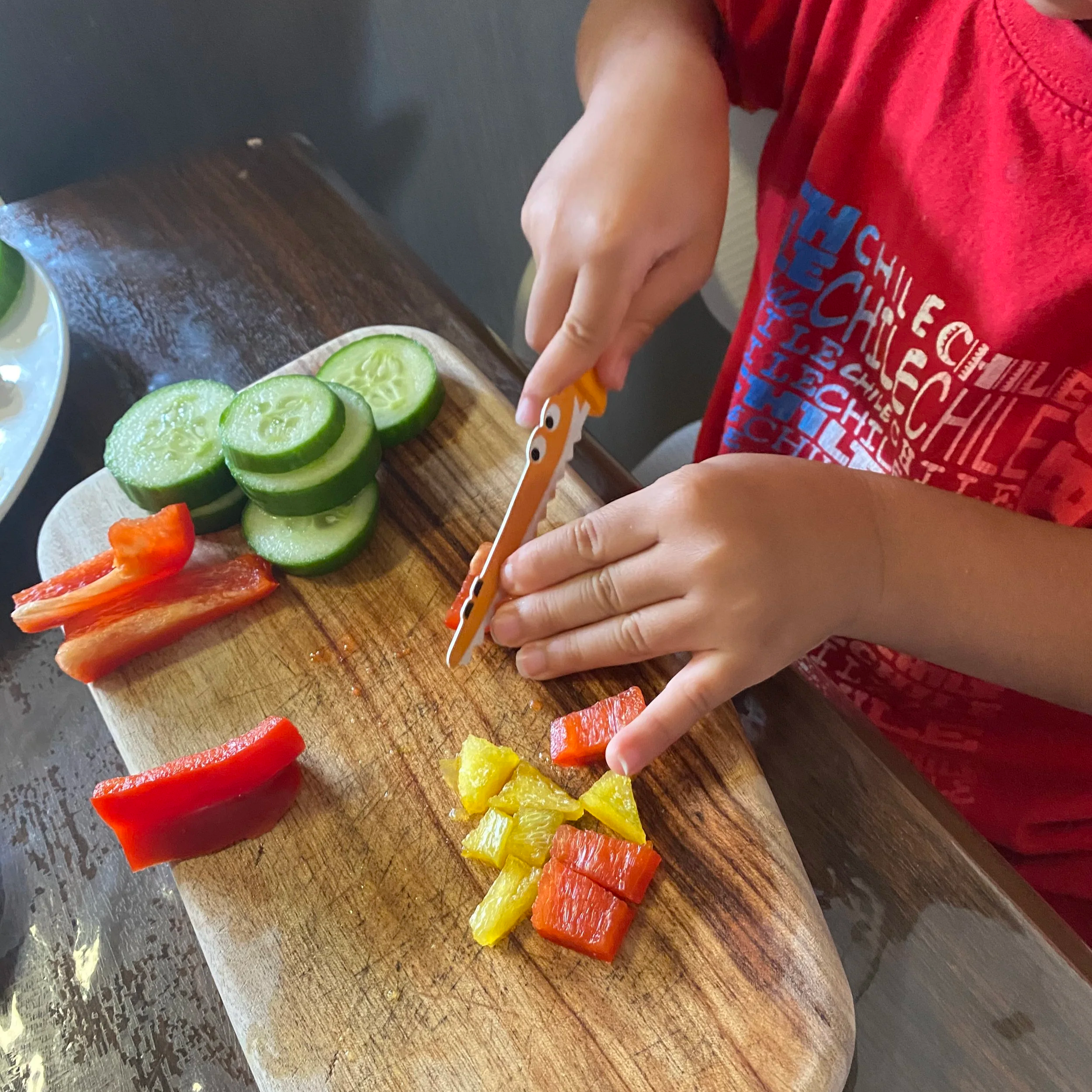 Child slicing colorful vegetables, including yellow, red, and orange bell peppers, on a wooden cutting board with sliced cucumber and red bell pepper nearby.