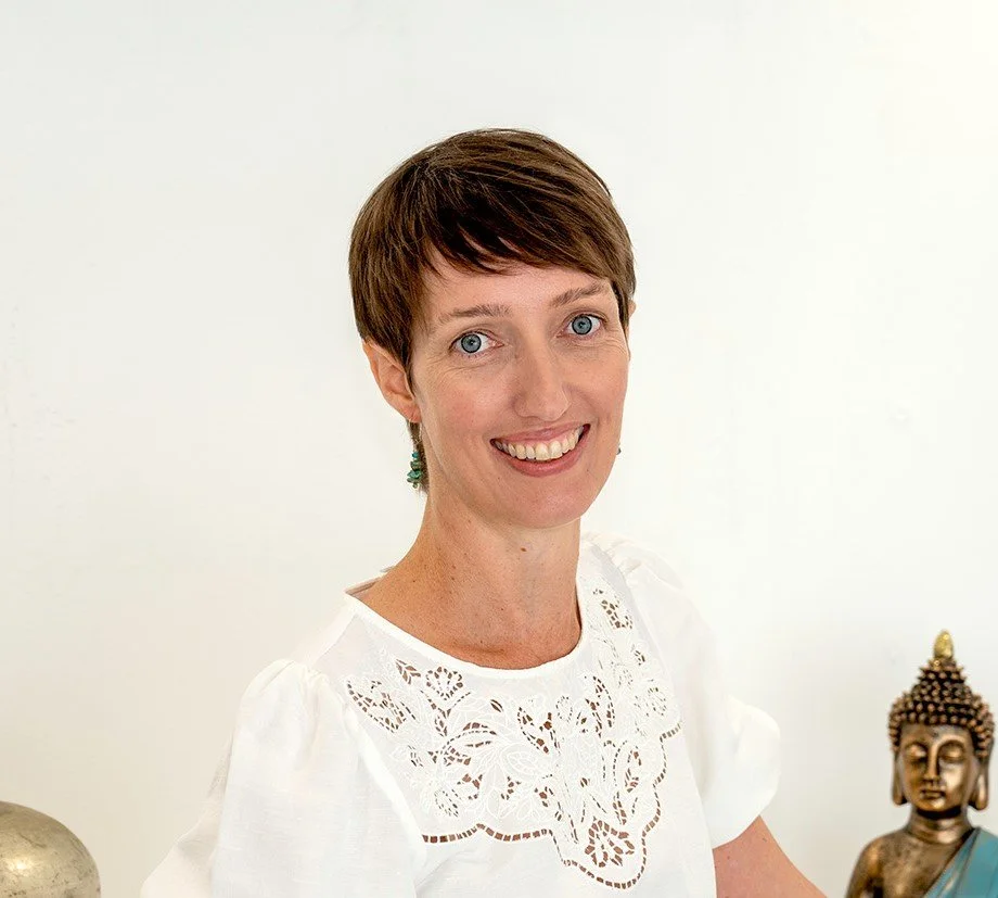 A woman with short brown hair and blue eyes smiling in front of a plain white wall, wearing a white embroidered top, with a small part of a Buddha statue visible on her right side.