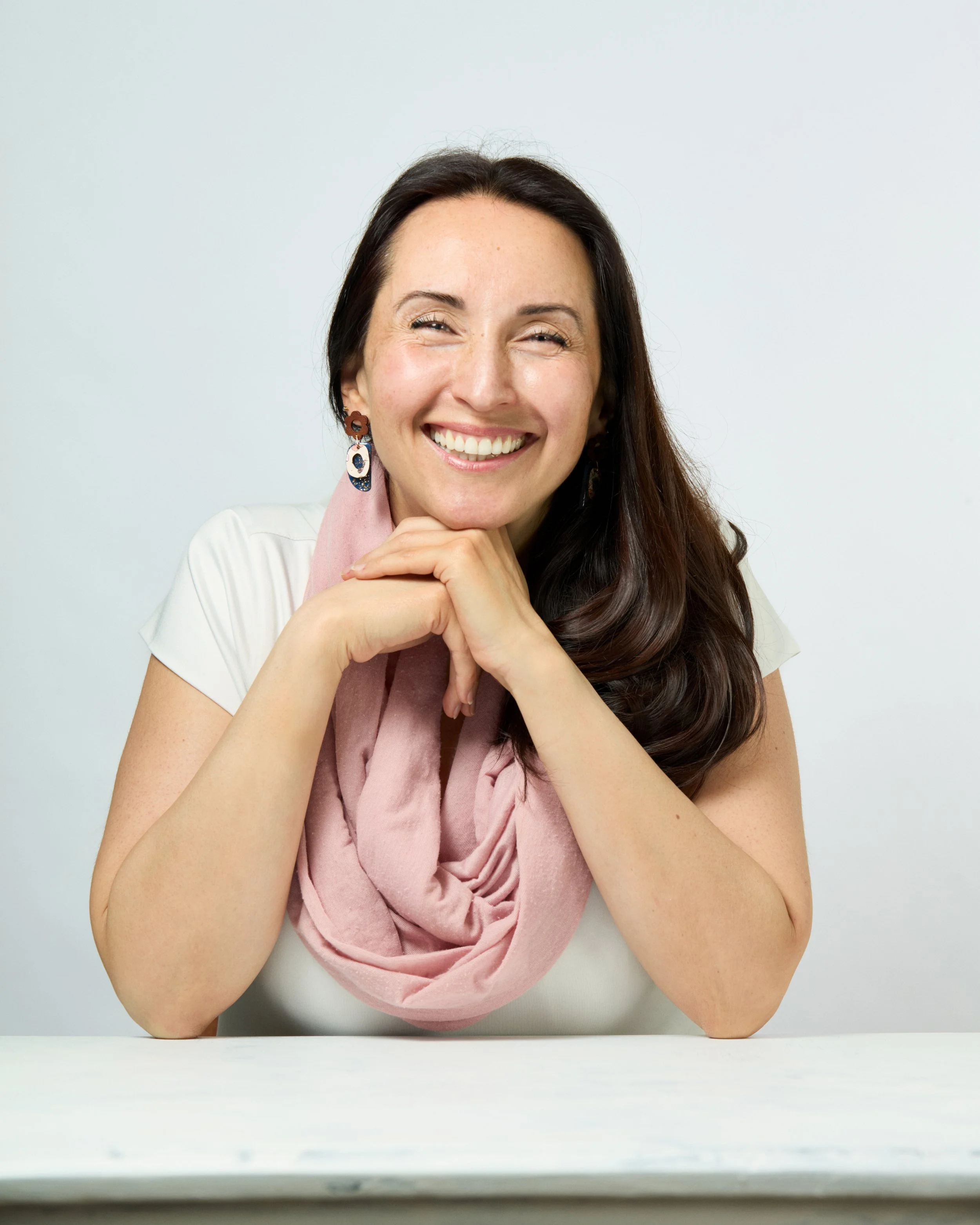 A woman with long dark hair smiling, resting her chin on her hands, wearing earrings, a white blouse, and a pink scarf, sitting at a white table against a plain light background.