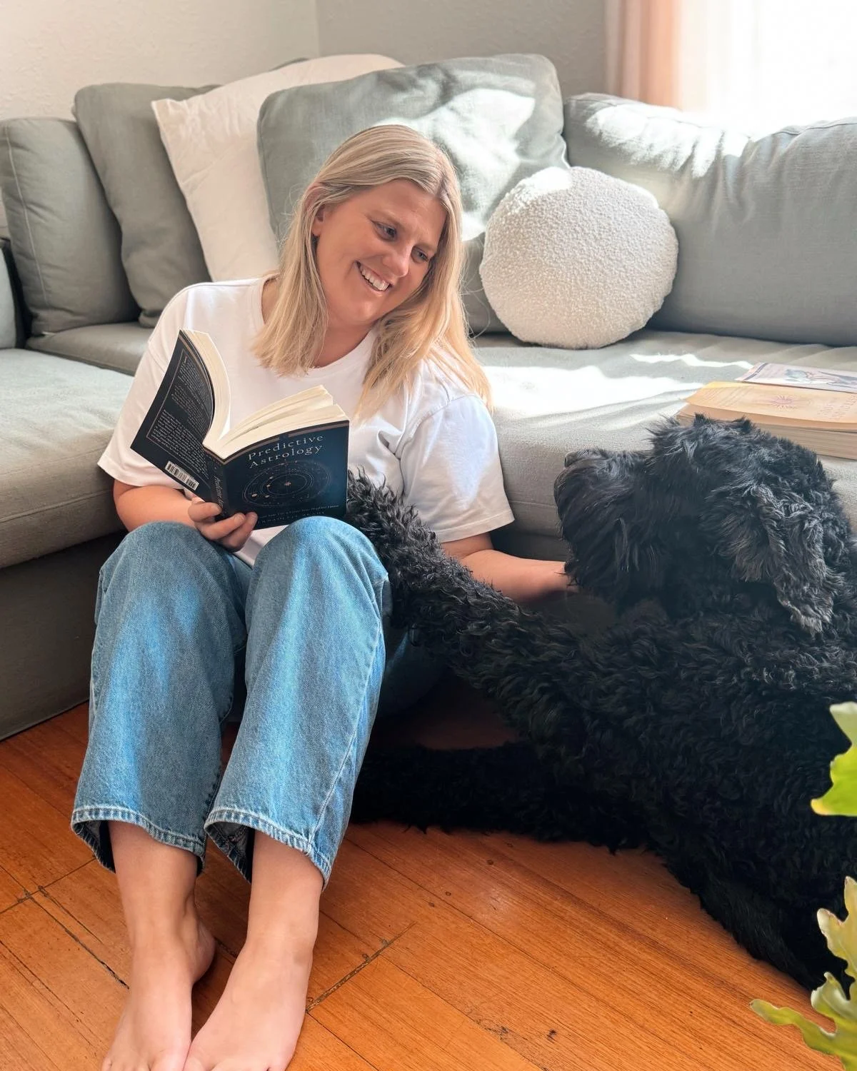 A woman sitting on the floor with her back against a couch, reading a book titled 'Predictive Astrology'. She is smiling and petting a large black dog lying next to her. There are books on the floor and cushions on the couch.