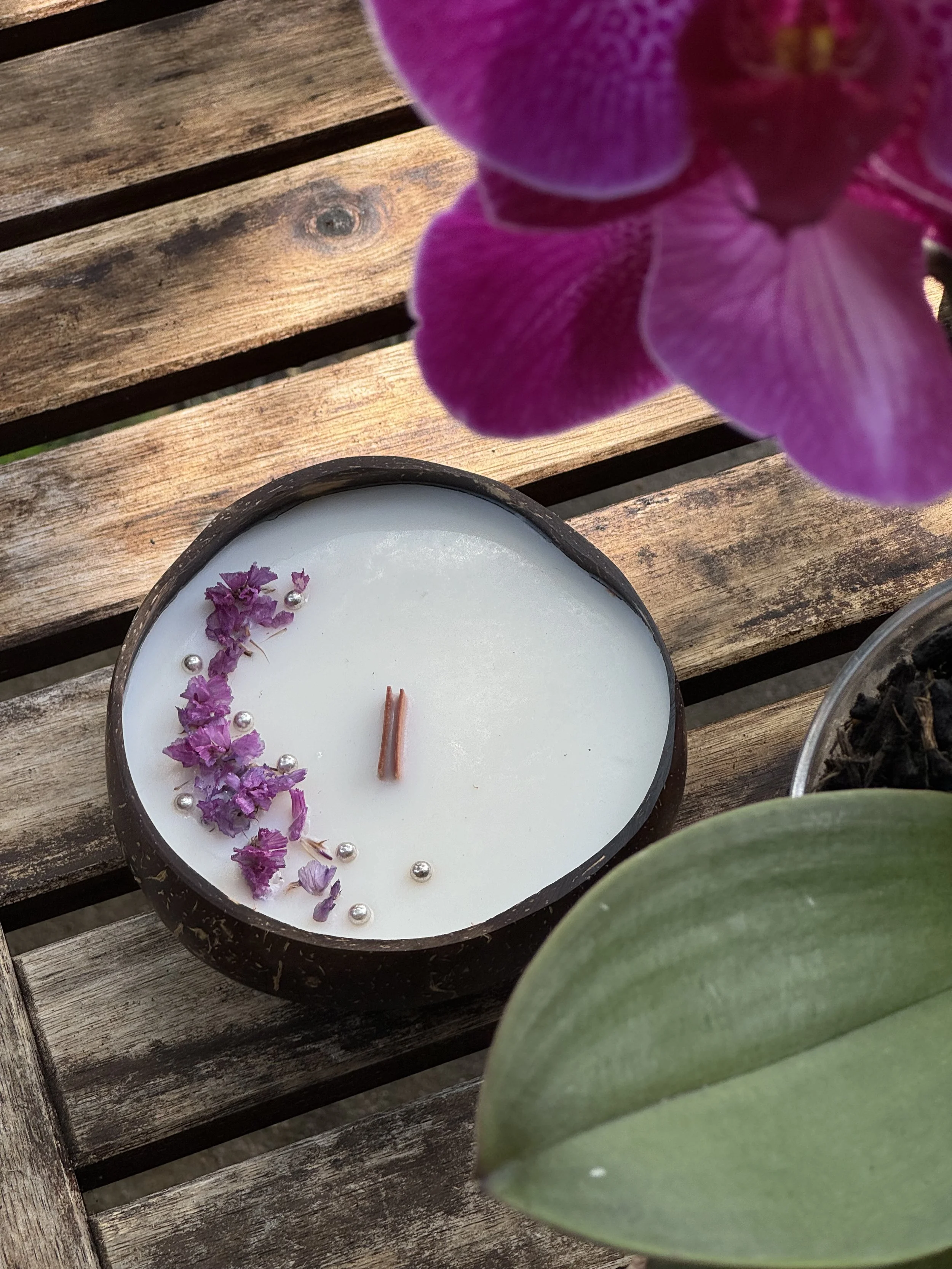 A white candle decorated with purple flowers and silver beads in a black holder, placed on a wooden surface. A purple orchid flower and a green leaf are partially visible in the foreground.