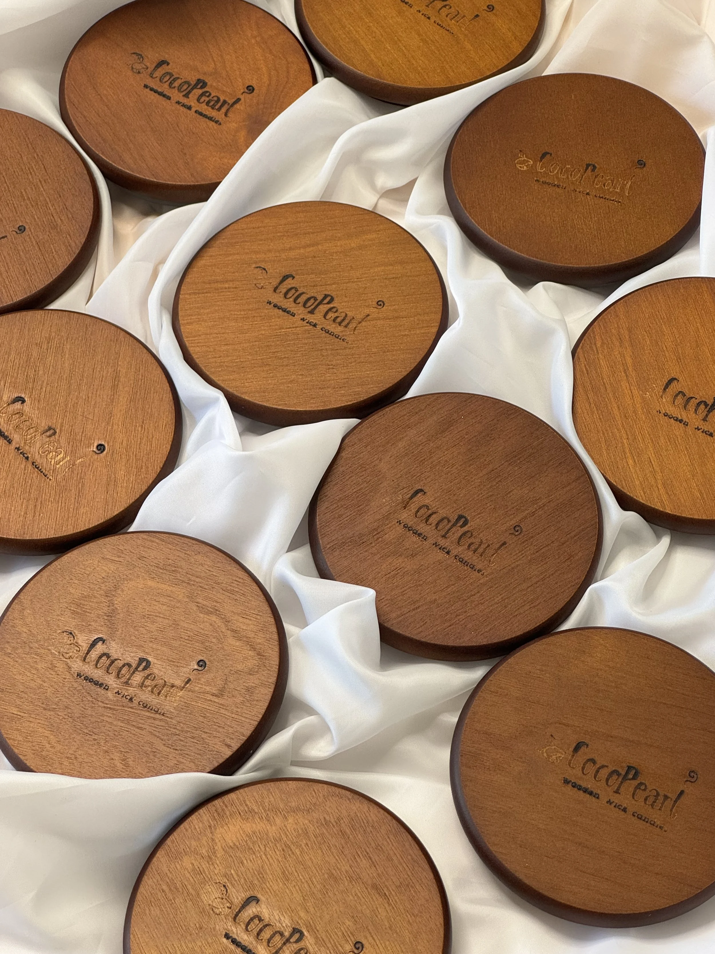 Multiple round wooden containers with the label 'CocoPearl' and the tagline 'wooded wick candles' placed on a white, silky fabric.