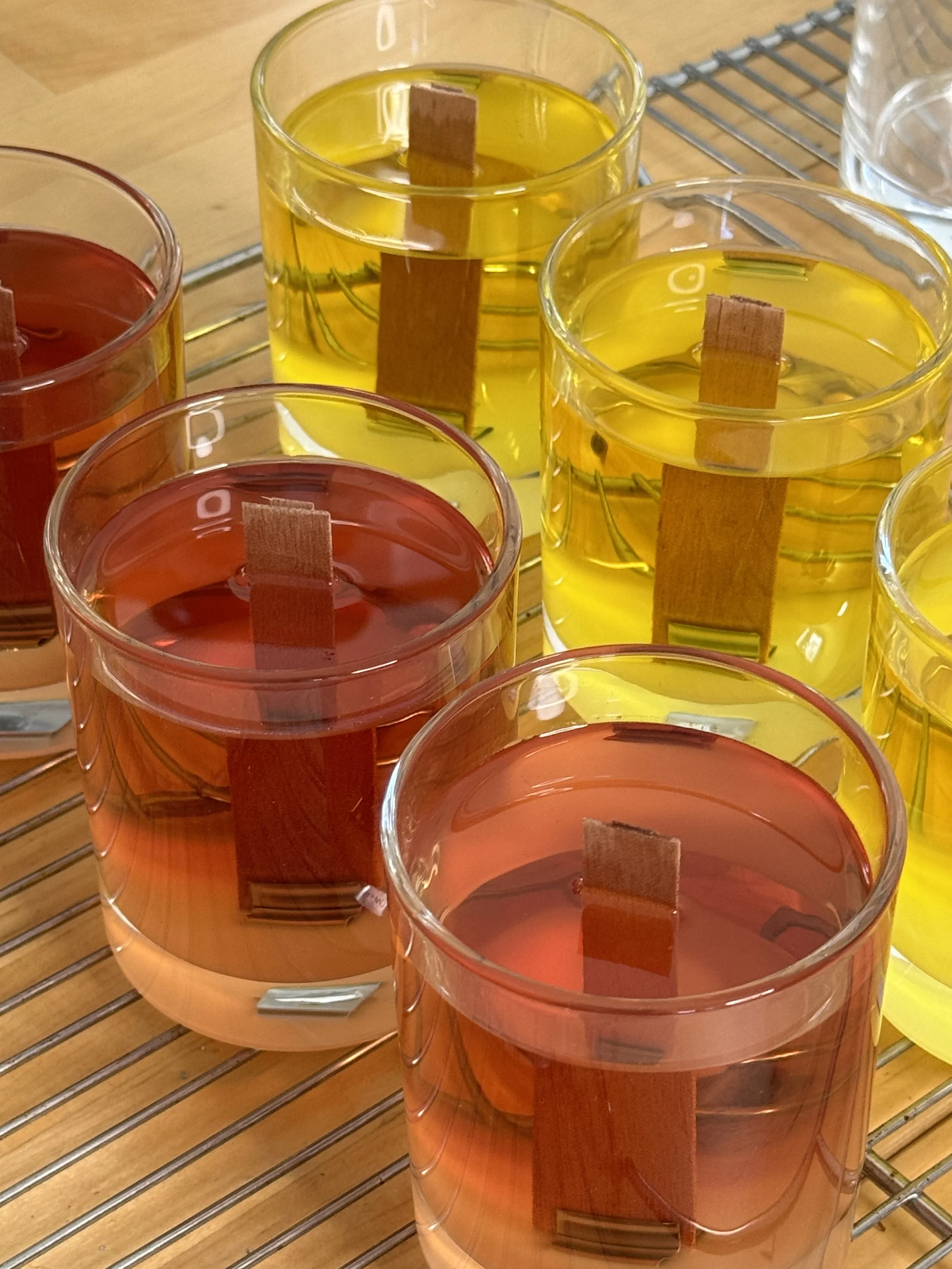 Colorful candles in clear glass holders with wooden sticks, placed on a metal cooling rack on a wooden surface.