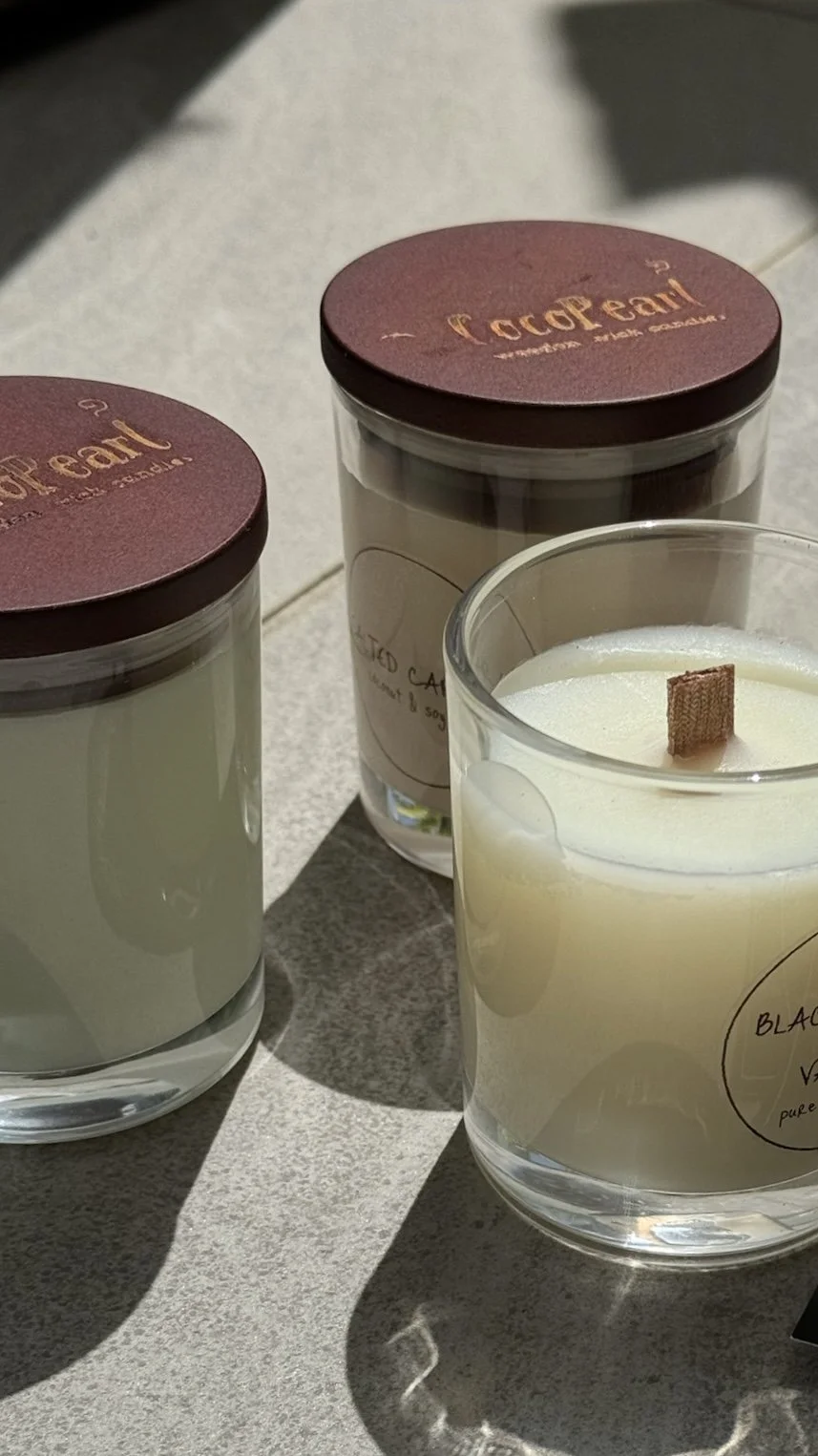 Three candles in glass containers with labeled wax, two on the left with brown lids, and one on the right with a wooden wick, placed on a gray surface with shadows.