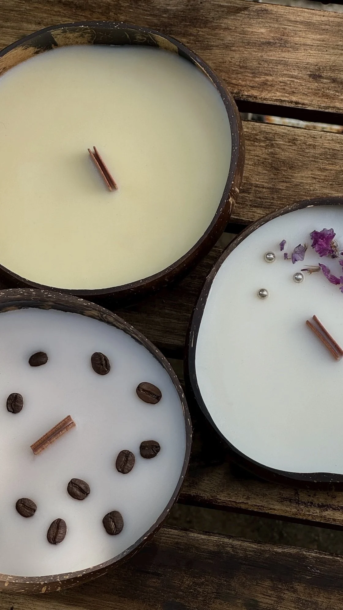 Three coffee-scented candles in dark containers on a wooden surface, decorated with coffee beans, dried flowers, small silver beads, and cinnamon sticks.